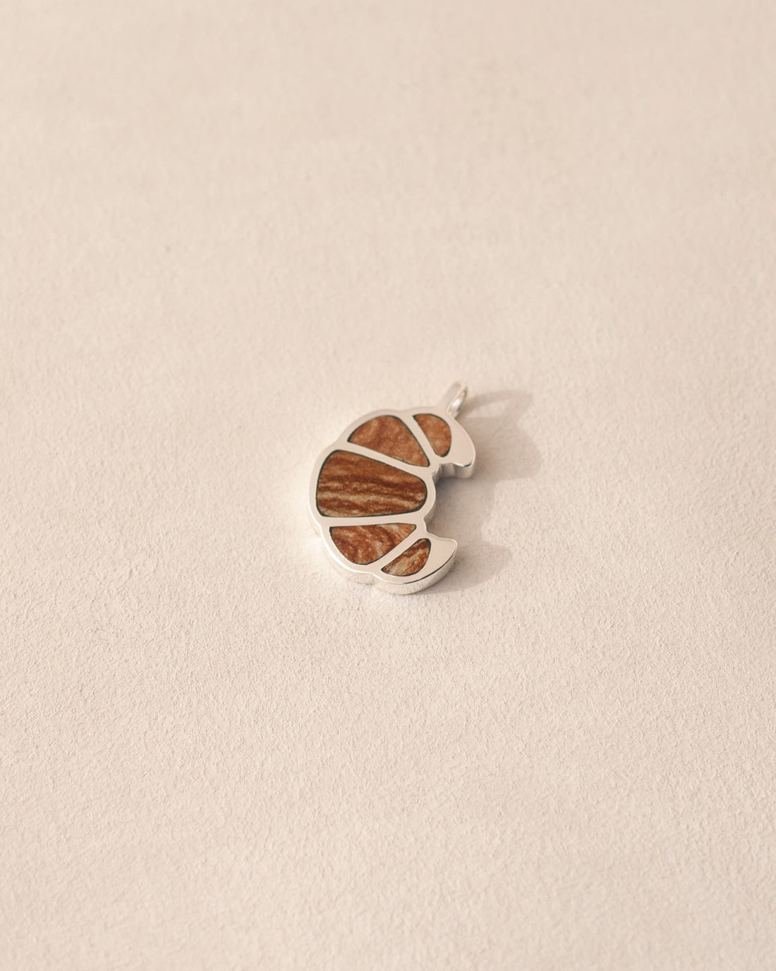 A silver charm in the shape of a croissant with flaky jasper inlaid into its design, displayed against a neutral background.