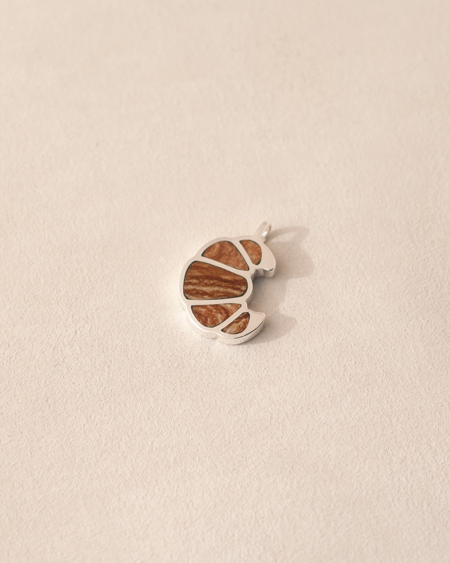 A silver charm in the shape of a croissant with flaky jasper inlaid into its design, displayed against a neutral background.