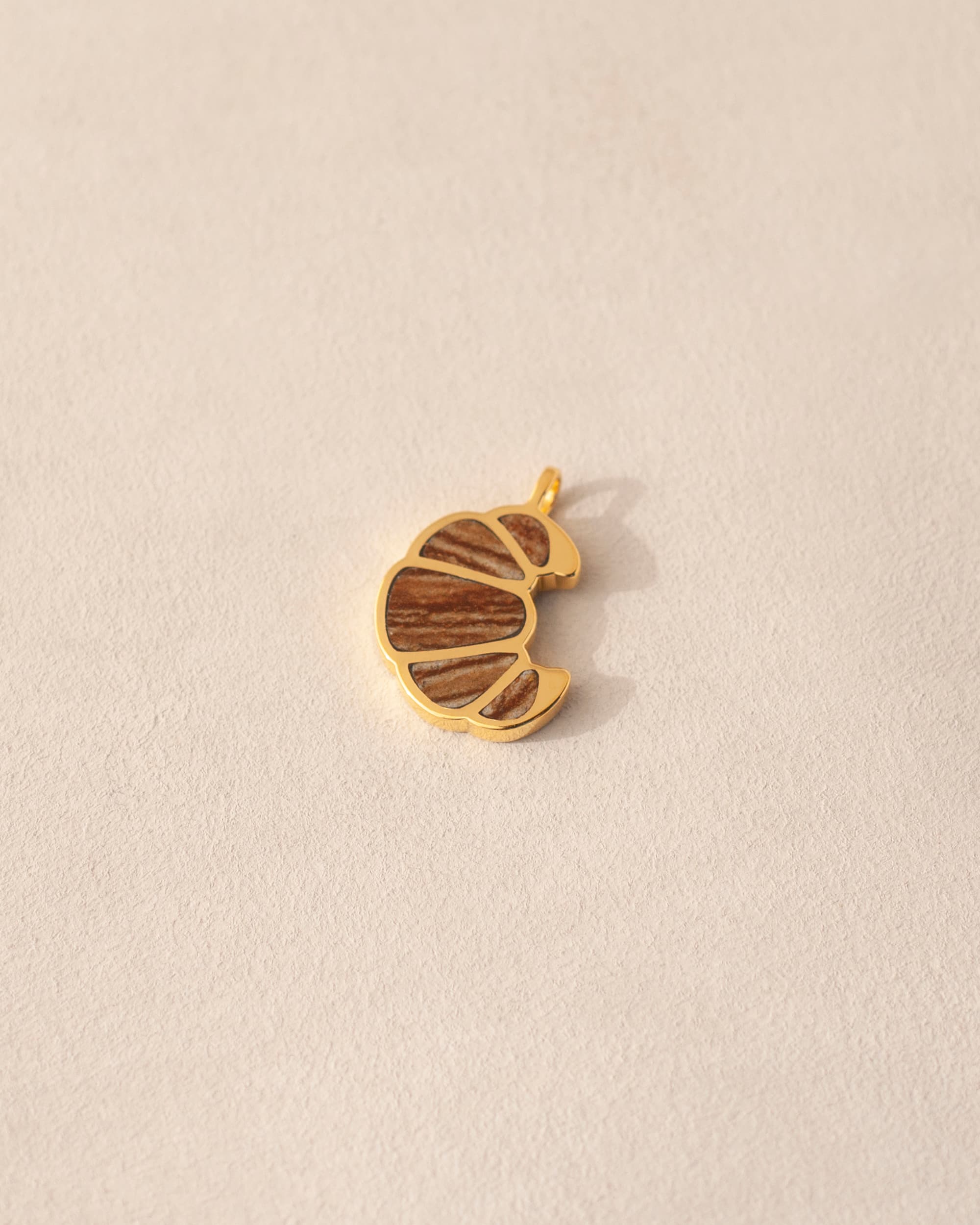 A gold-colored charm in the shape of a croissant, inlaid with jasper, placed on a neutral background.