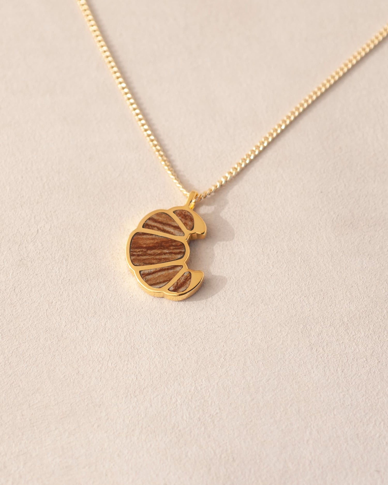 a gold charm shaped like a croissant, inlaid with jasper, dangling from a gold chain