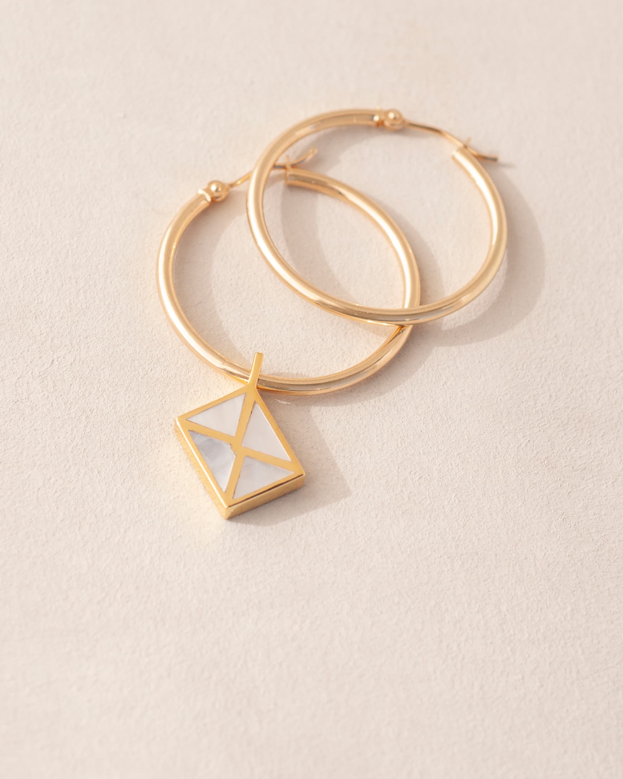 A gold charm shaped like an envelope dangling from gold hoops, on a light background.