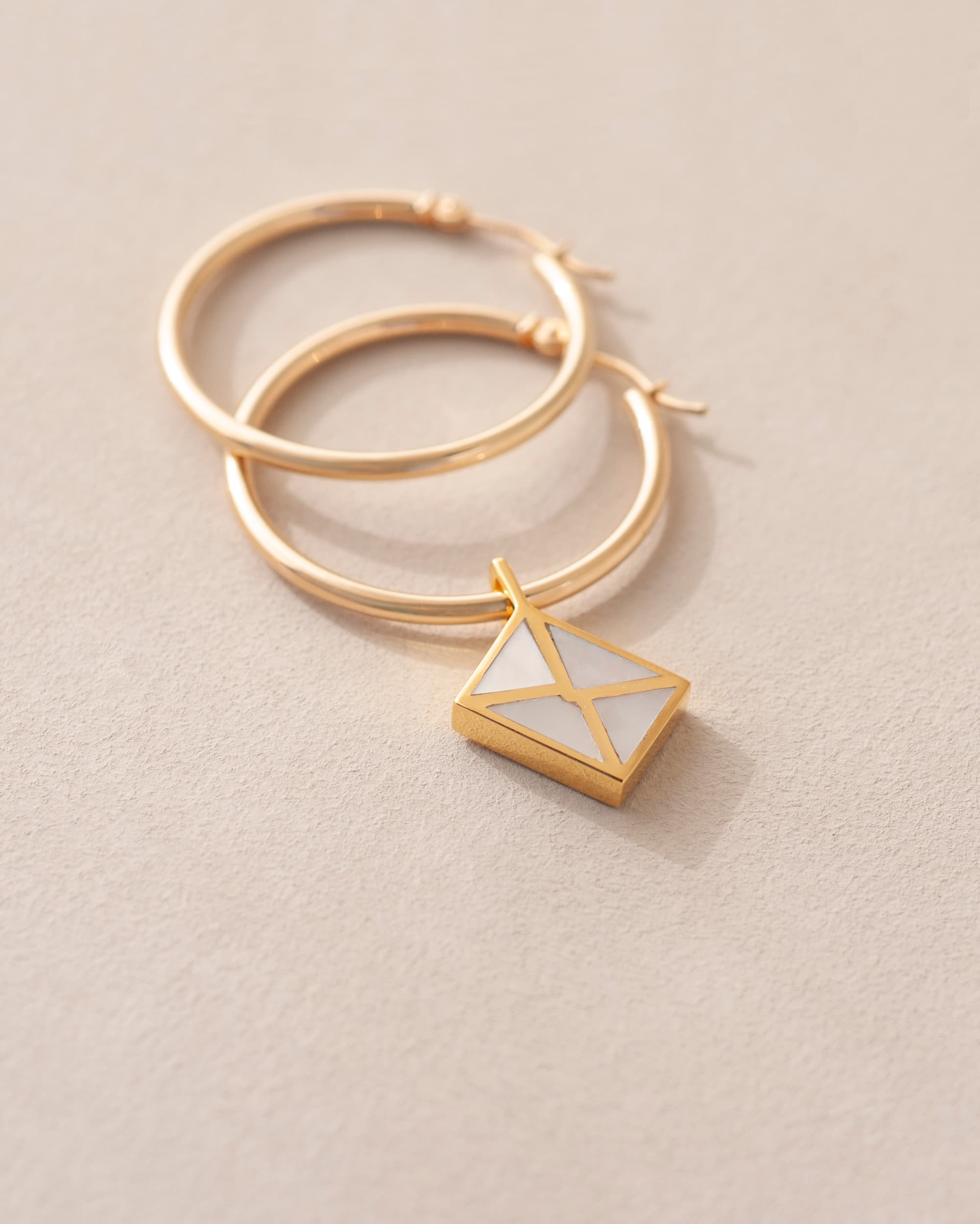 A gold charm shaped like an envelope dangling from gold hoops, on a light background.