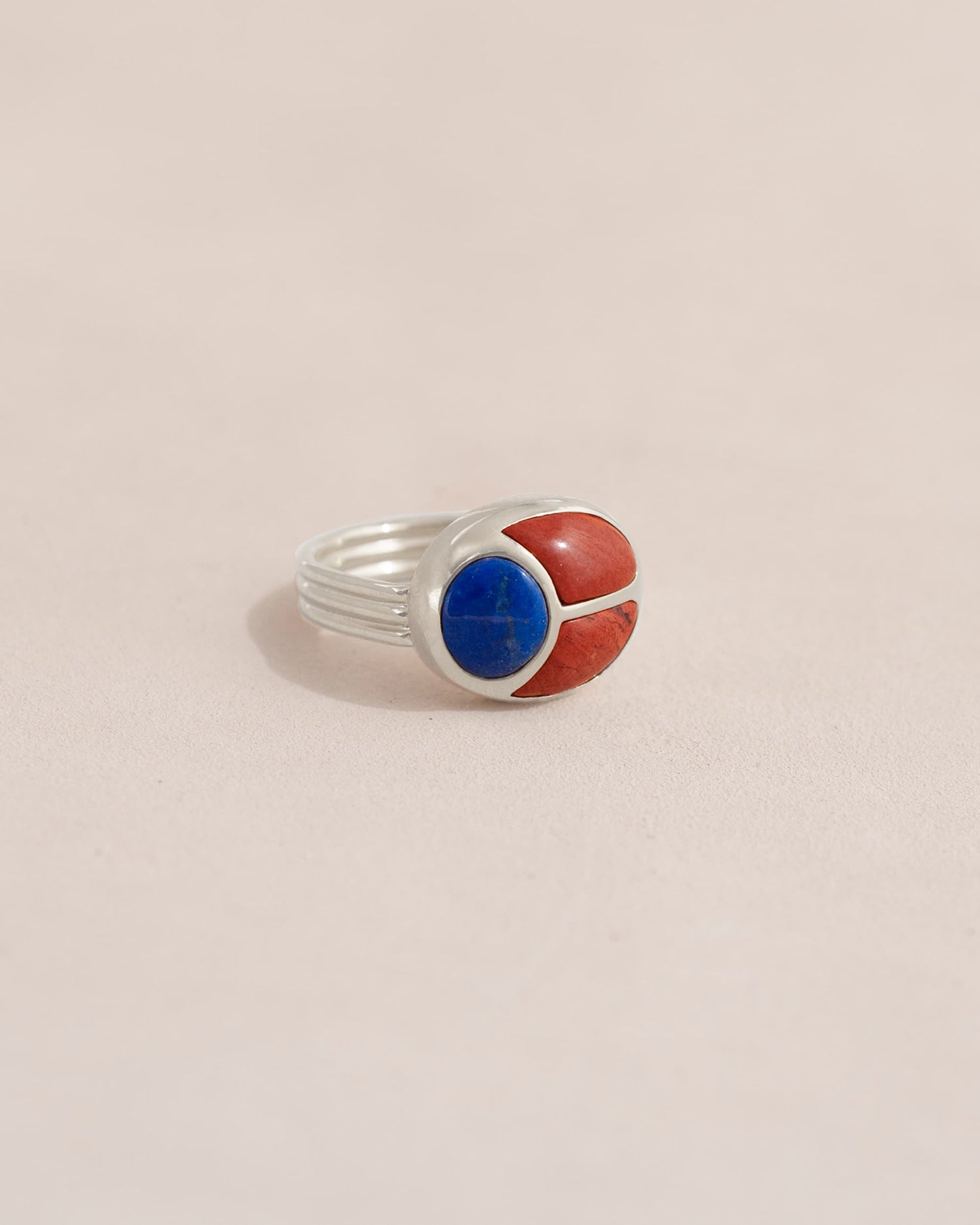 A silver ring featuring a scarab design with lapis lazuli and red jasper inlays, displayed against a neutral background.