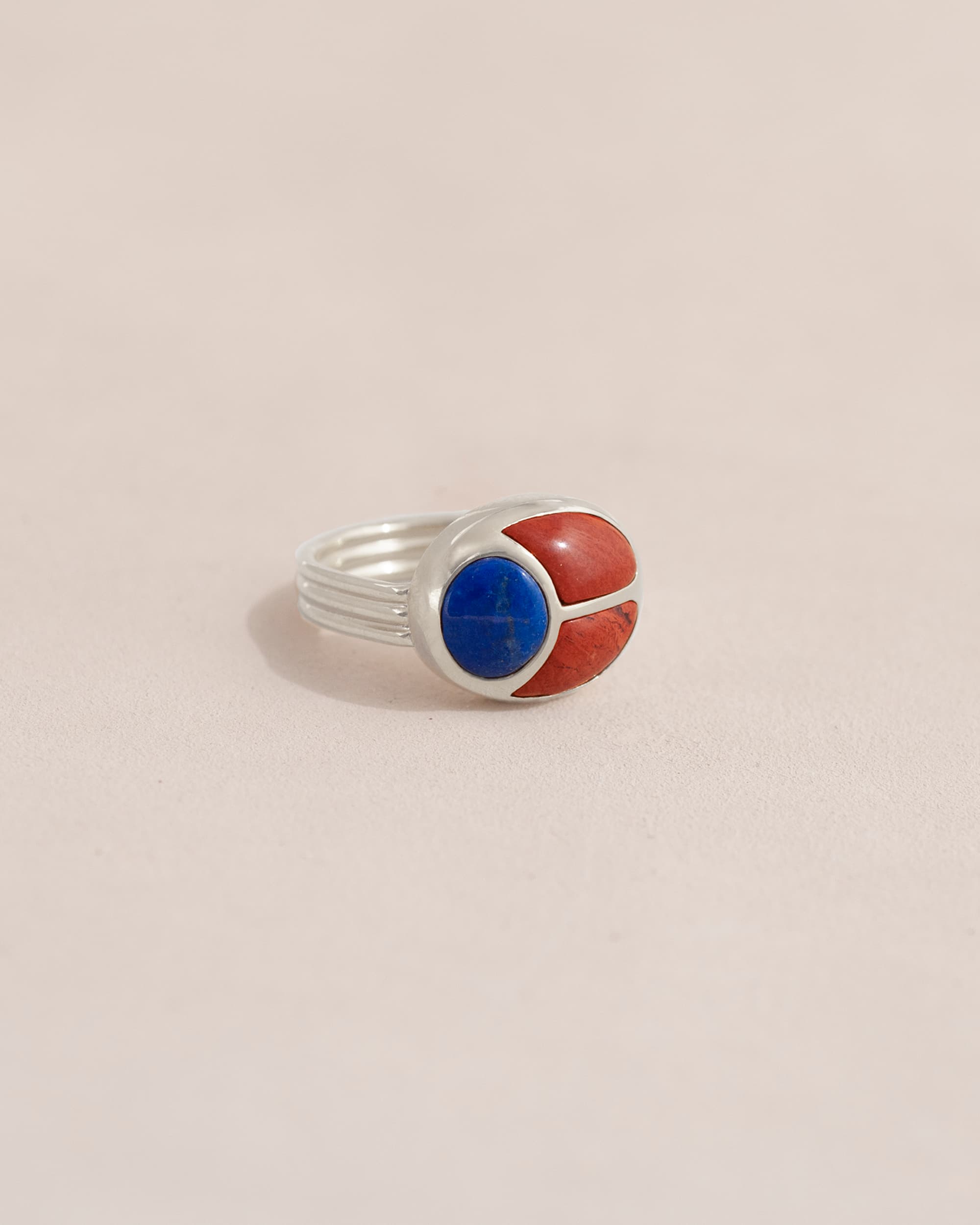 A silver ring featuring a scarab design with lapis lazuli and red jasper inlays, displayed against a neutral background.