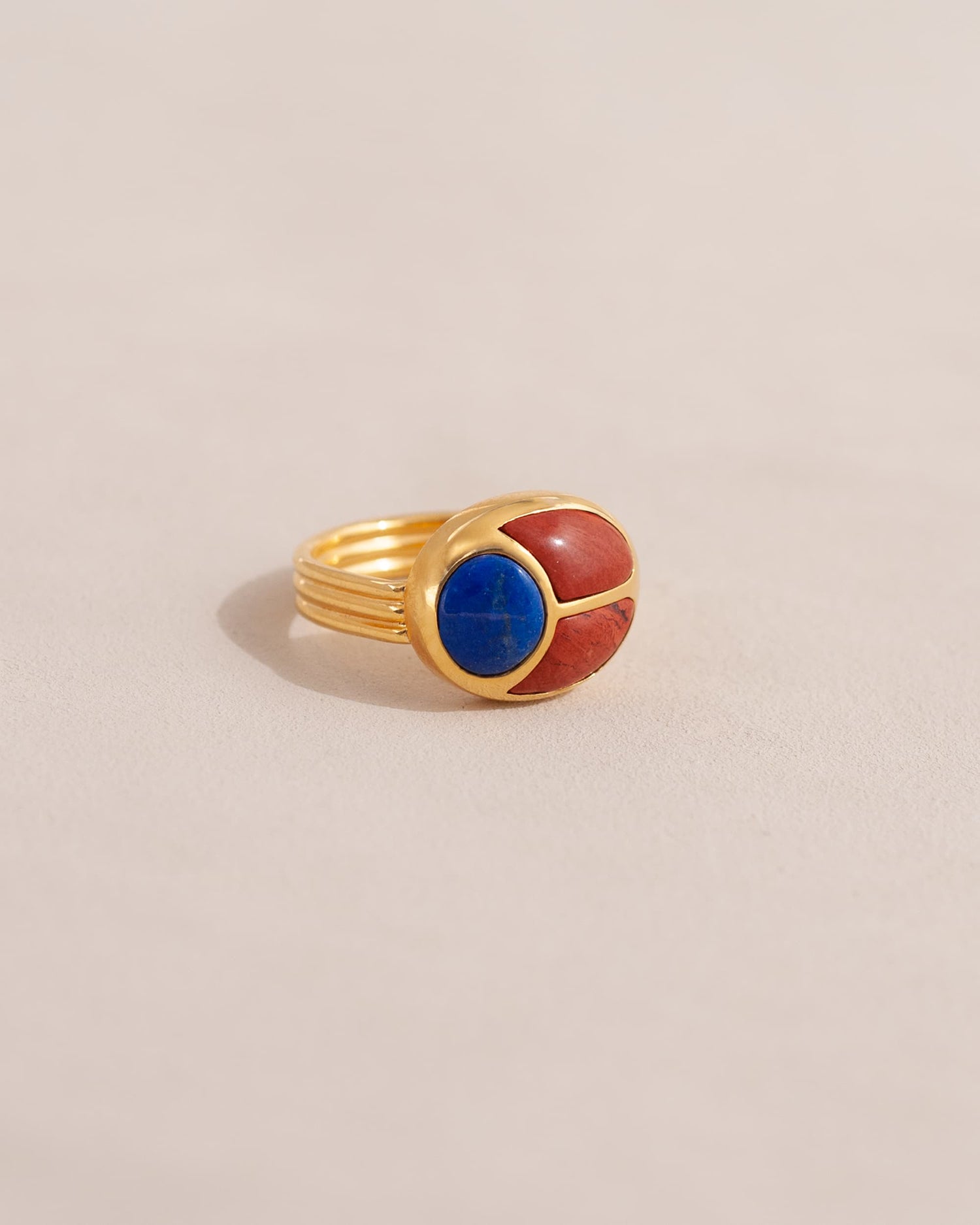 A gold ring featuring a scarab design with lapis lazuli and red jasper inlays, displayed against a neutral background.