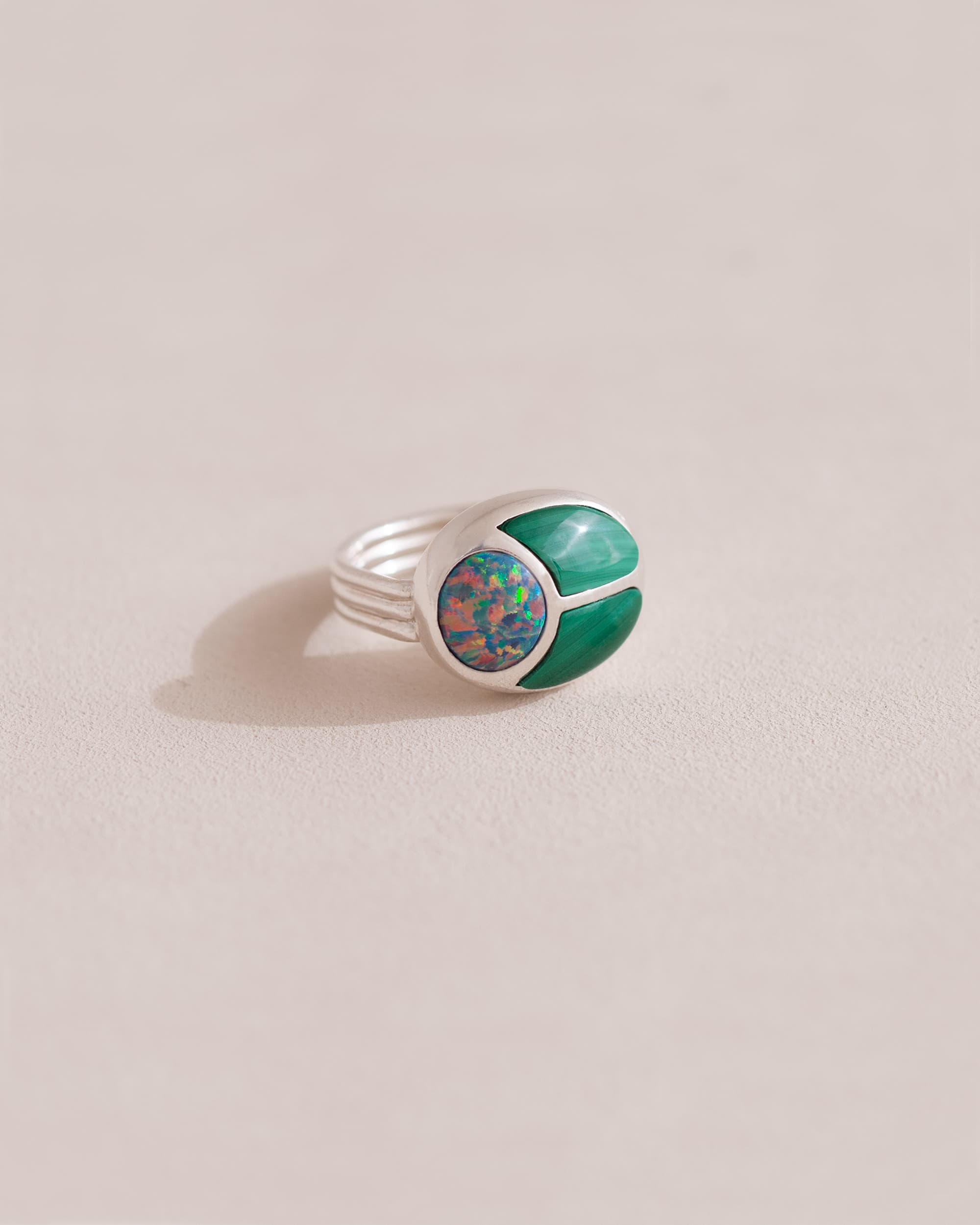 A silver ring featuring a central opal stone flanked by malachite stones on either side, set against a neutral background.