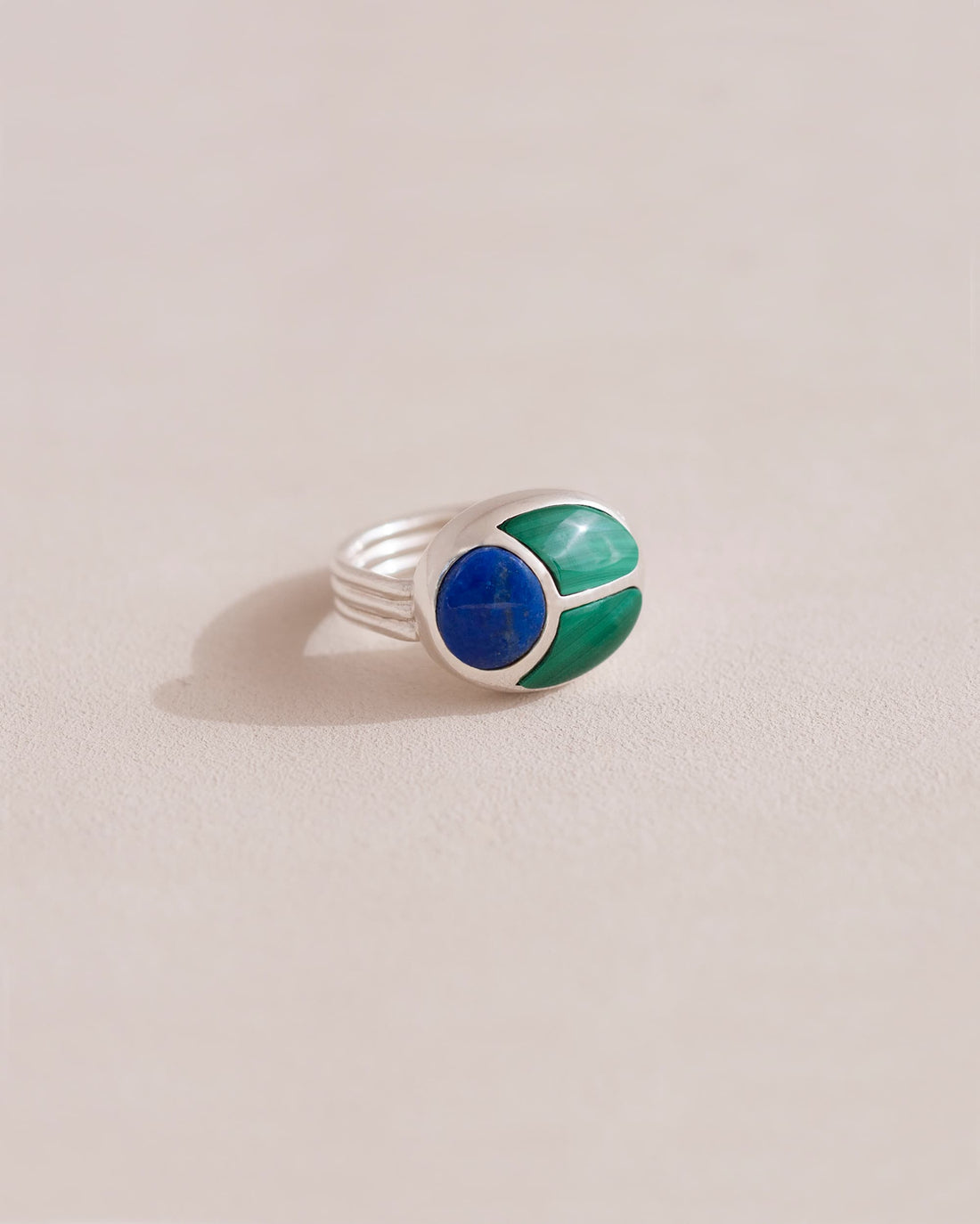 A silver ring featuring a central lapis lazuli stone flanked by malachite stones on either side, set against a neutral background.