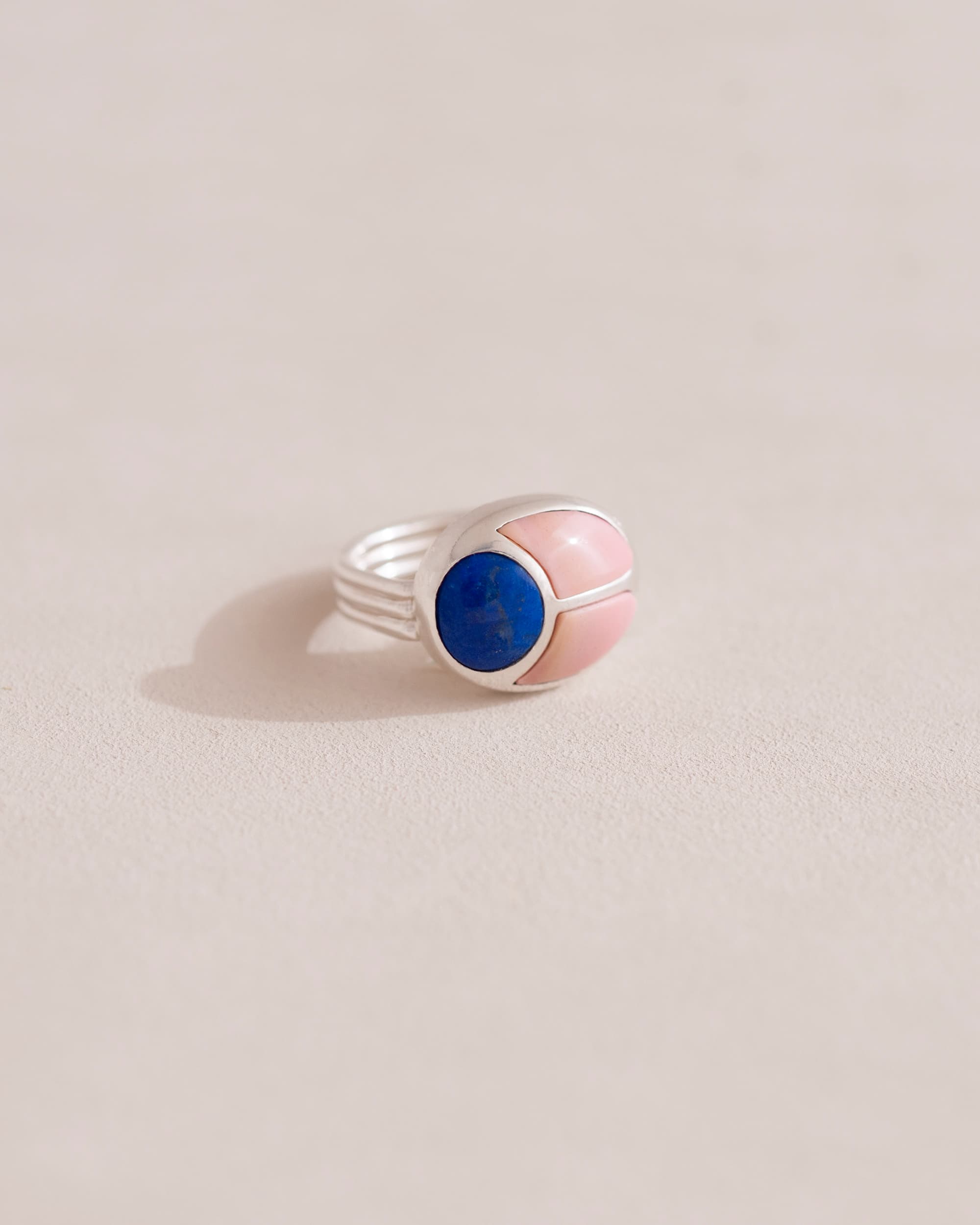 A silver ring featuring a central lapis lazuli stone flanked by pink opal stones on either side, set against a neutral background.
