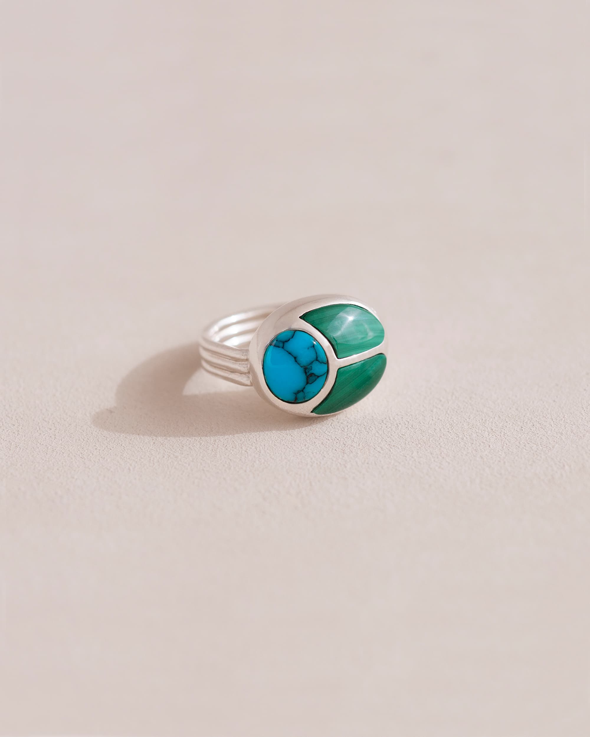 A silver ring featuring a central turquoise stone flanked by malachite stones on either side, set against a neutral background.