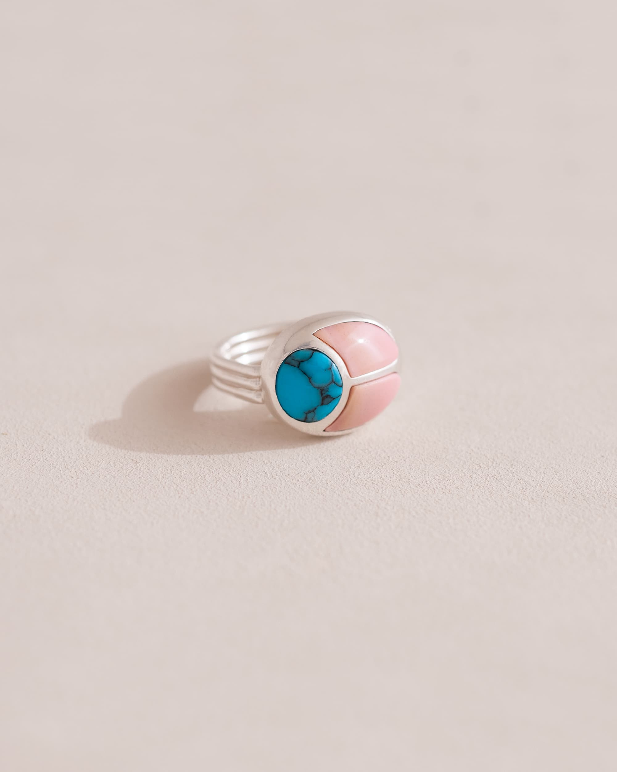 A silver ring featuring a central turquoise stone flanked by pink opal stones on either side, set against a neutral background.