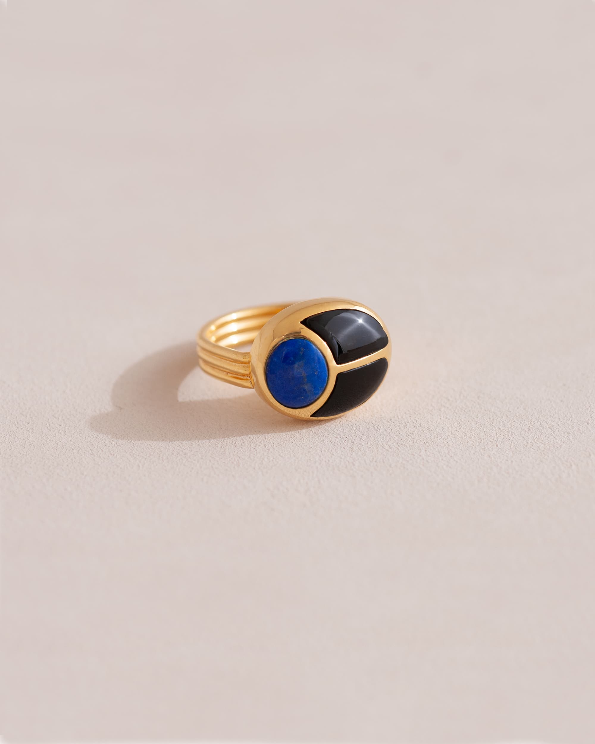 A gold ring featuring a central lapis lazuli stone flanked by onyx stones on either side, set against a neutral background.