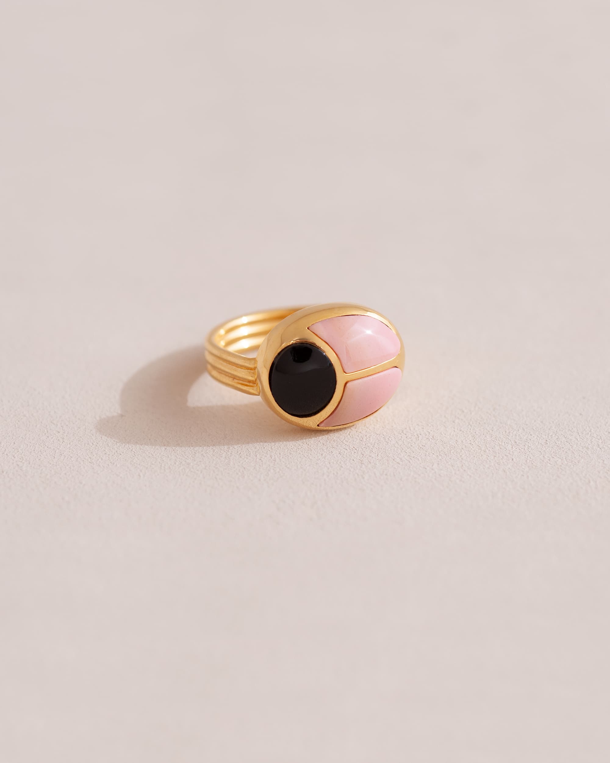 A gold ring featuring a central onyx stone flanked by pink opal stones on either side, set against a neutral background.