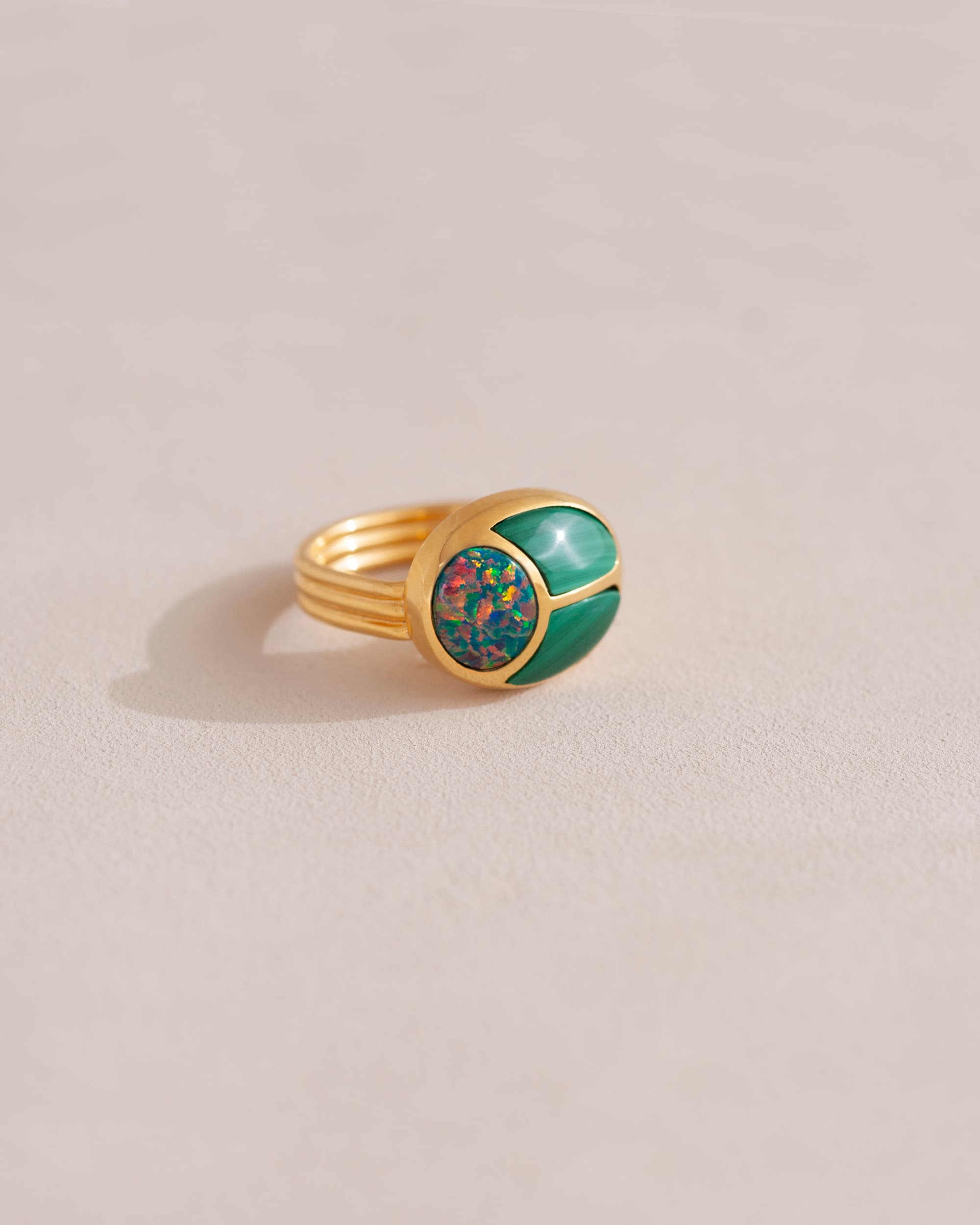 A gold ring featuring a central opal stone flanked by malachite stones on either side, set against a neutral background.