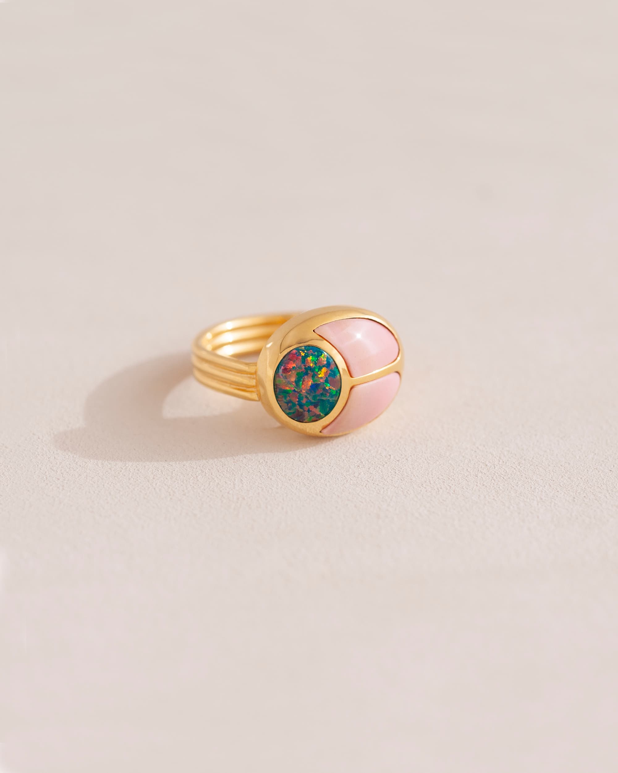 A gold ring featuring a central opal stone flanked by pink opal stones on either side, set against a neutral background.