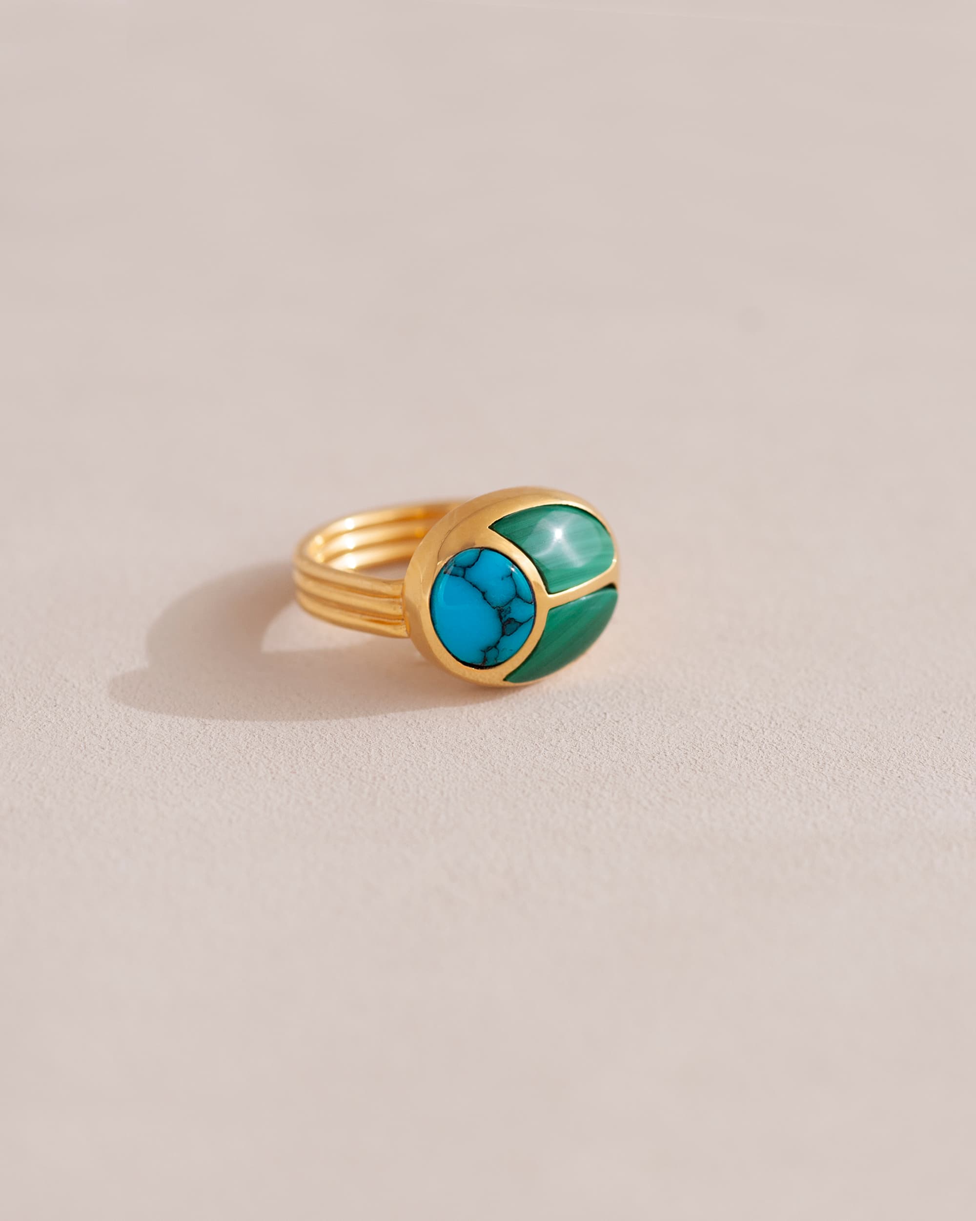 A gold ring featuring a central turquoise stone flanked by malachite stones on either side, set against a neutral background.