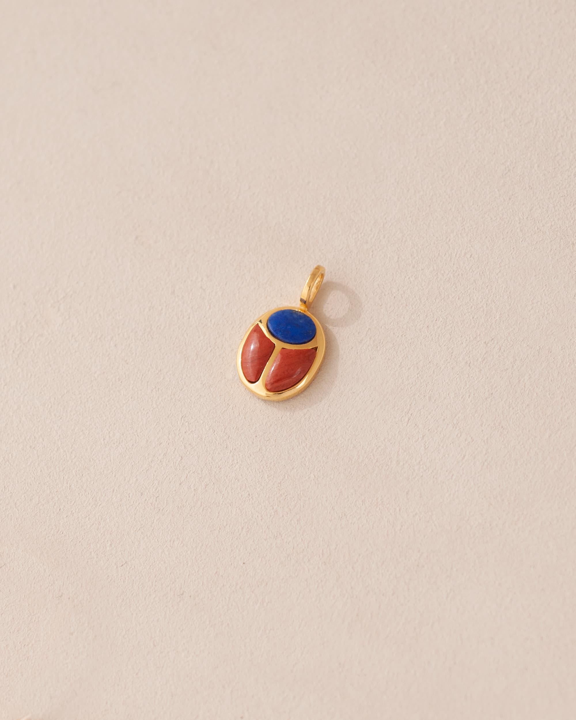 A gold charm featuring a scarab design with lapis lazuli and red jasper inlays, displayed against a neutral background.