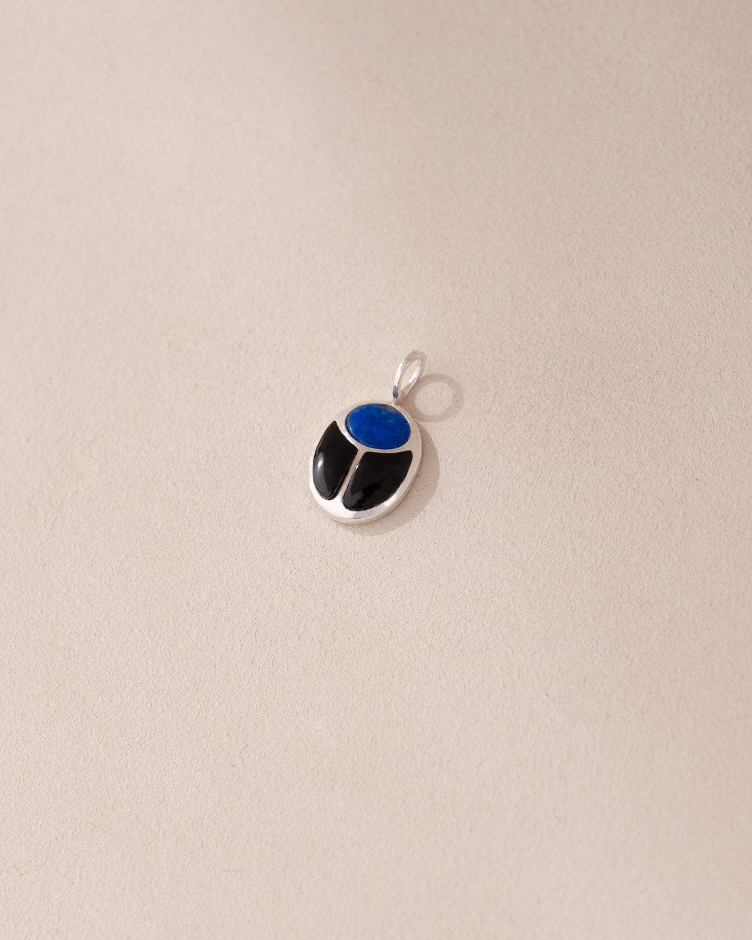 A silver-colored charm with a blue lapis lazuli center and black onyx surround, placed against a beige background.