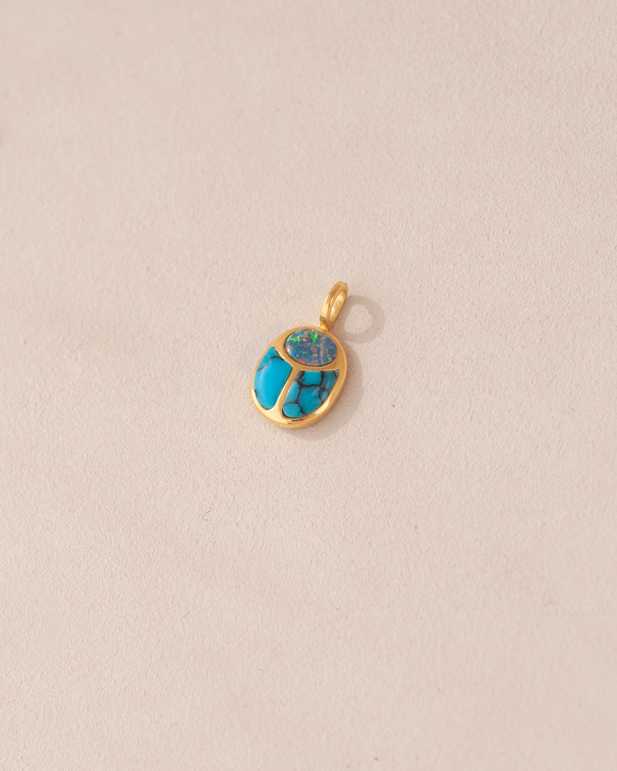 A gold charm featuring a scarab design with starry opal and turquoise inlay, placed against a beige background.