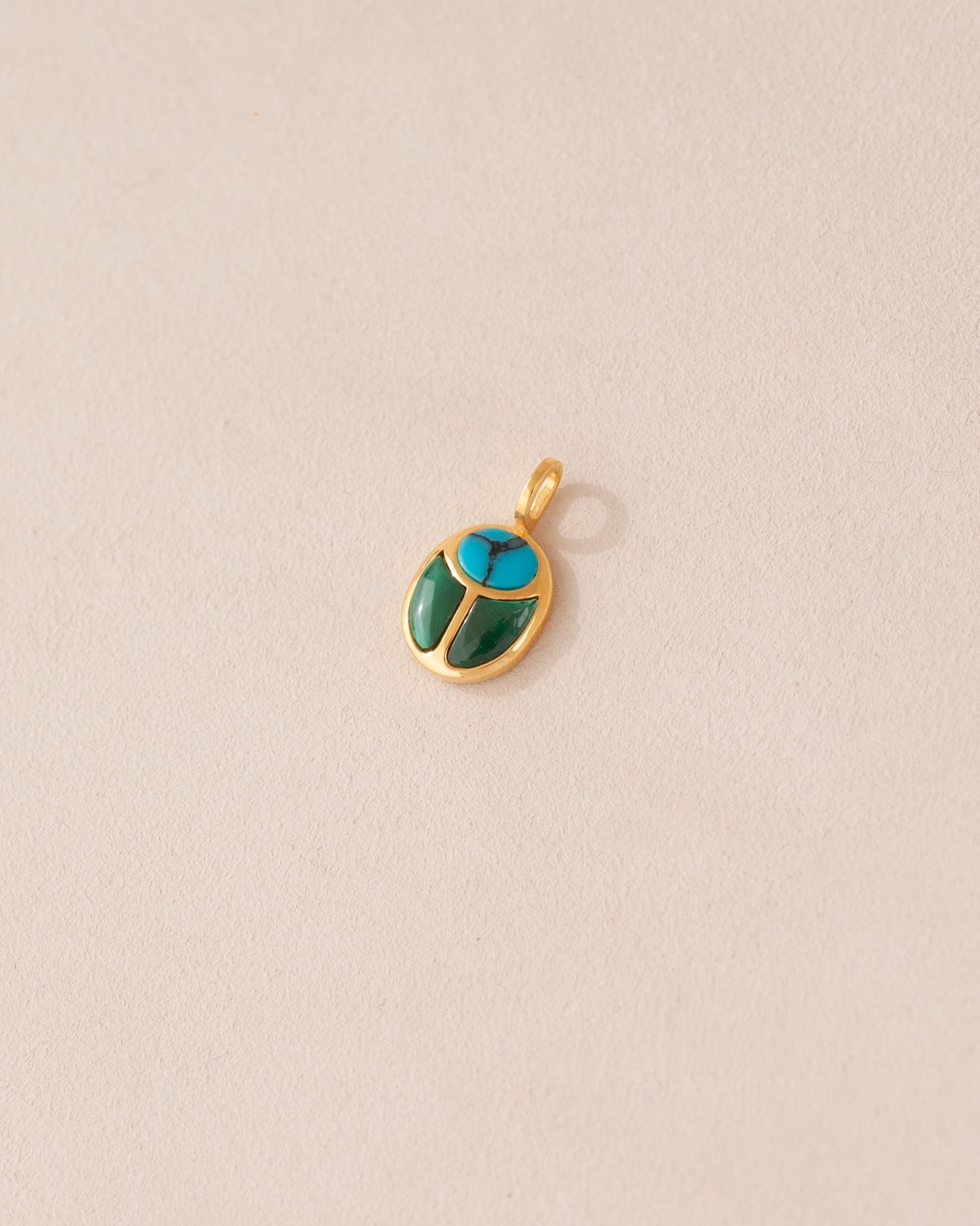 A small turquoise and malachite stone charm in a gold setting, shaped like a scarab, displayed against a light background.