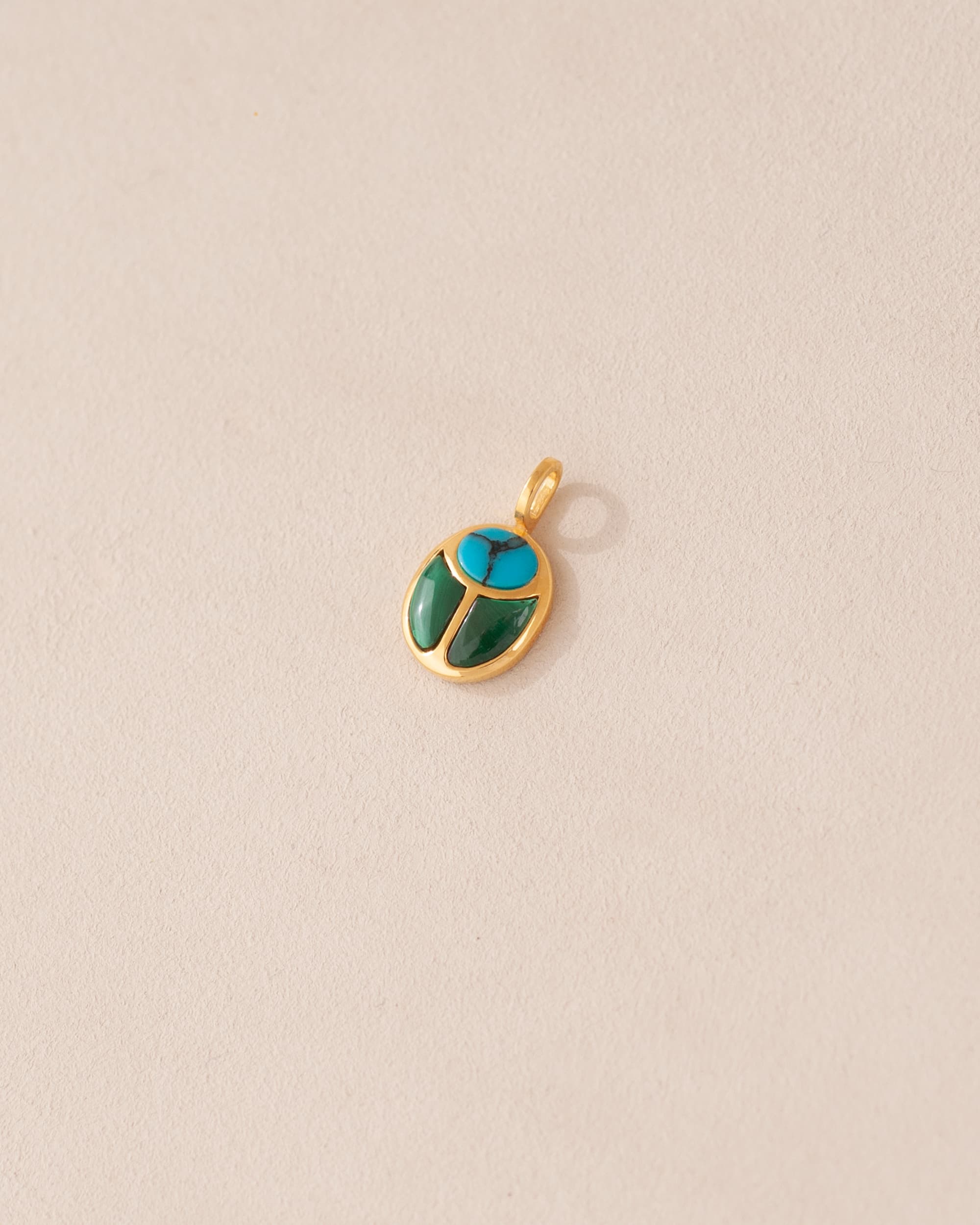 A small turquoise and malachite stone charm in a gold setting, shaped like a scarab, displayed against a light background.