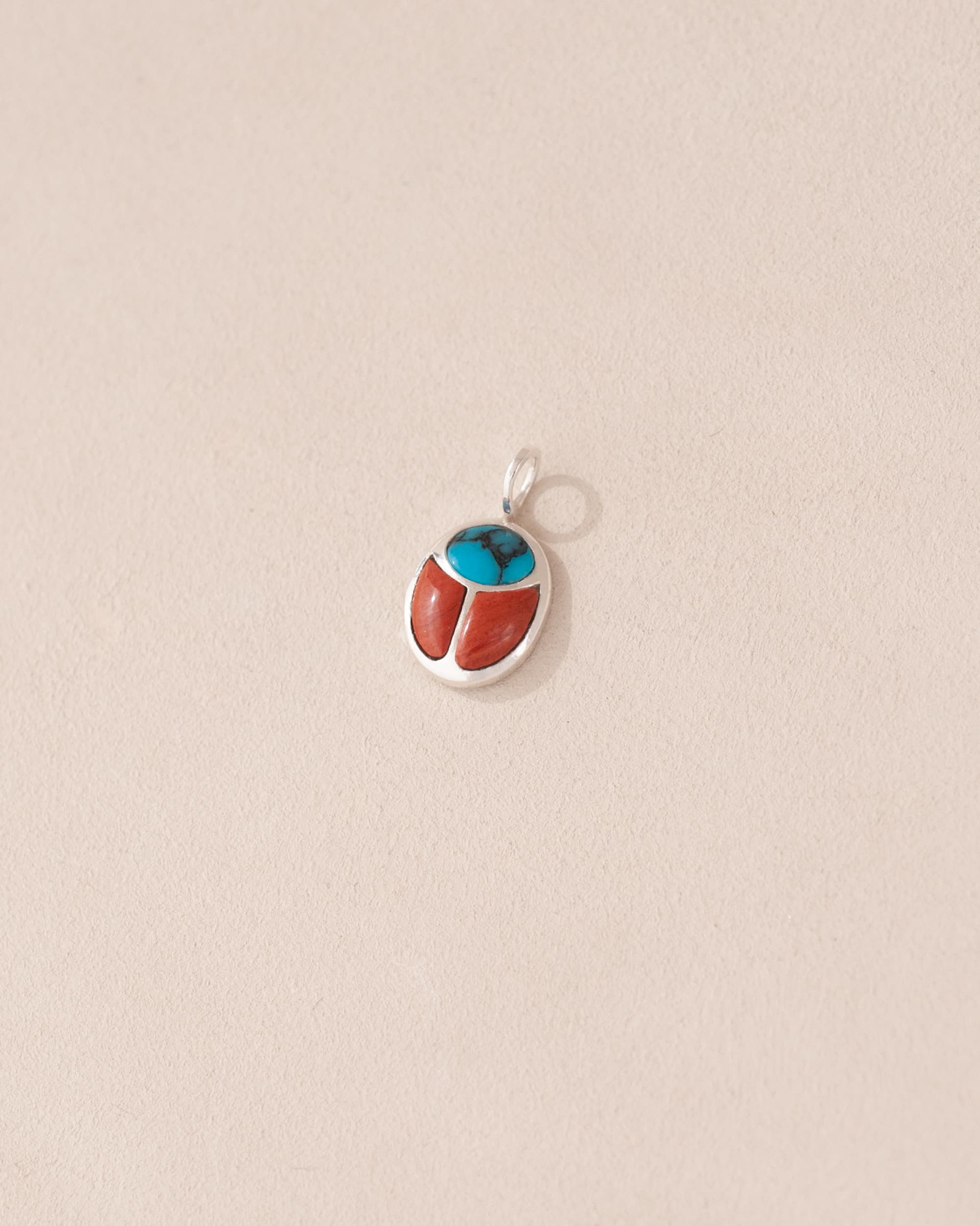A small silver charm featuring a turquoise and red jasper design, shaped like a scarab amulet.