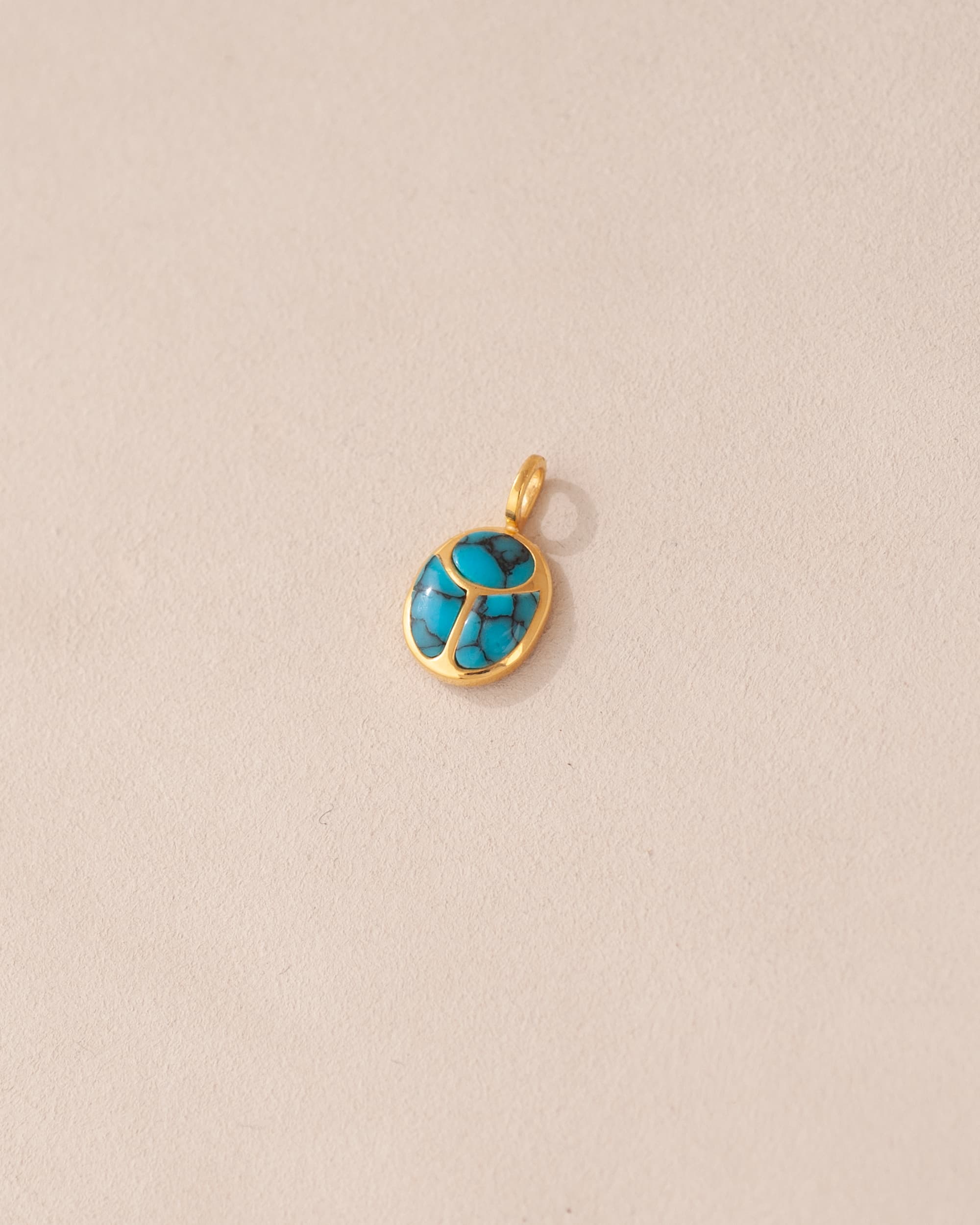A turquoise-colored scarab charm set in 14K yellow gold, placed against a beige background.