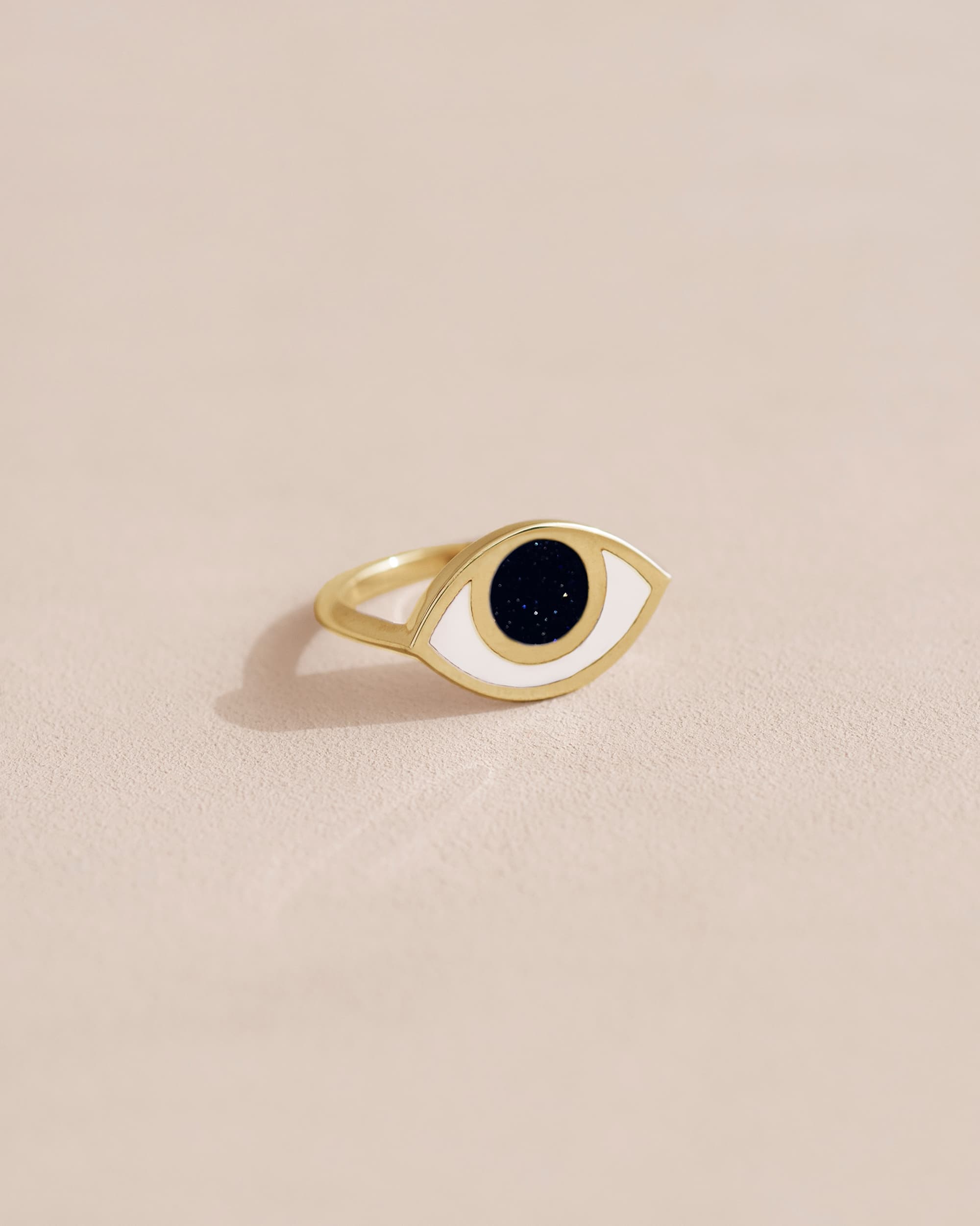 A gold-colored ring with a black stone shaped like an eye, placed on a neutral background.