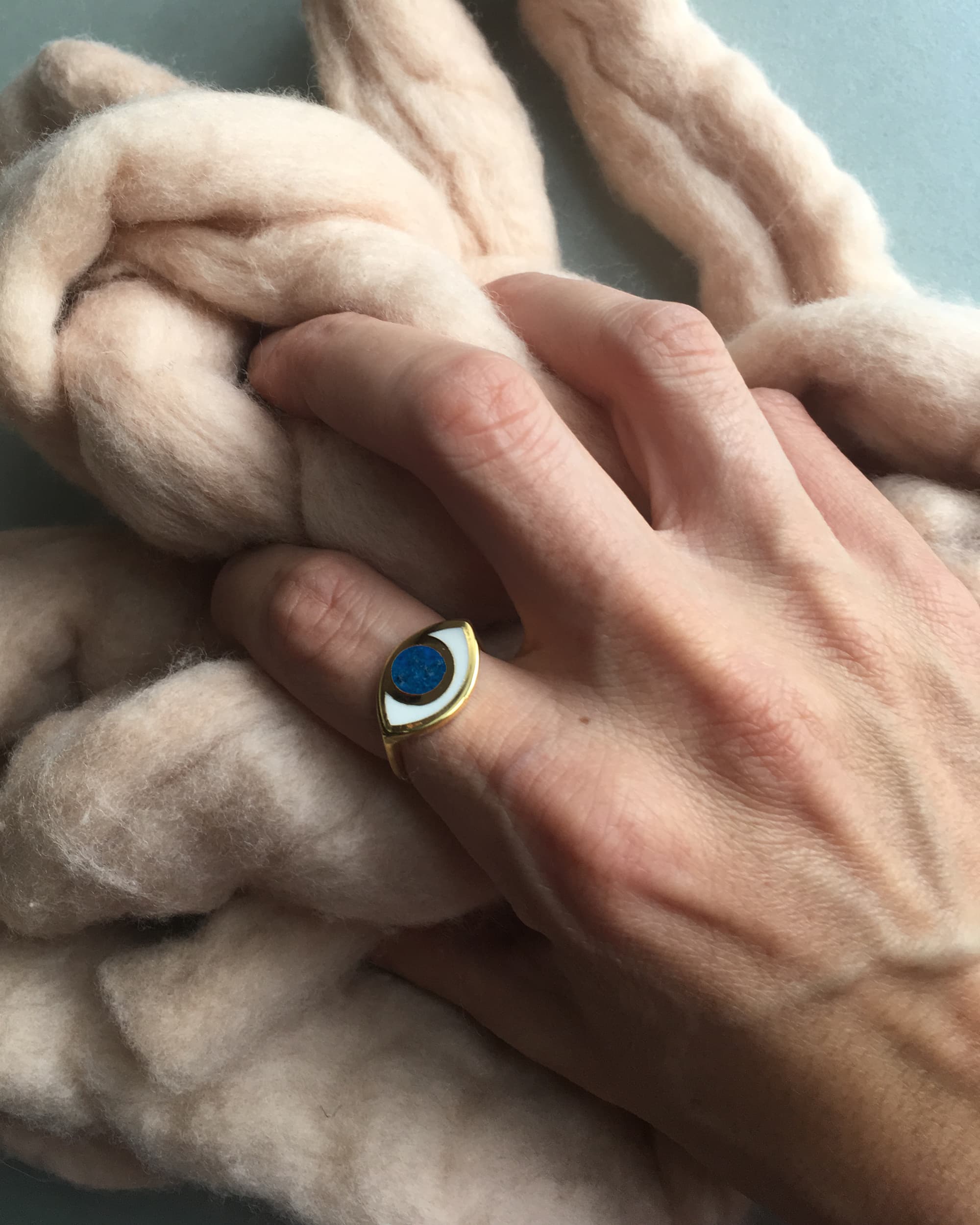 a hand wearing a gold eye shaped ring and holding pink wool
