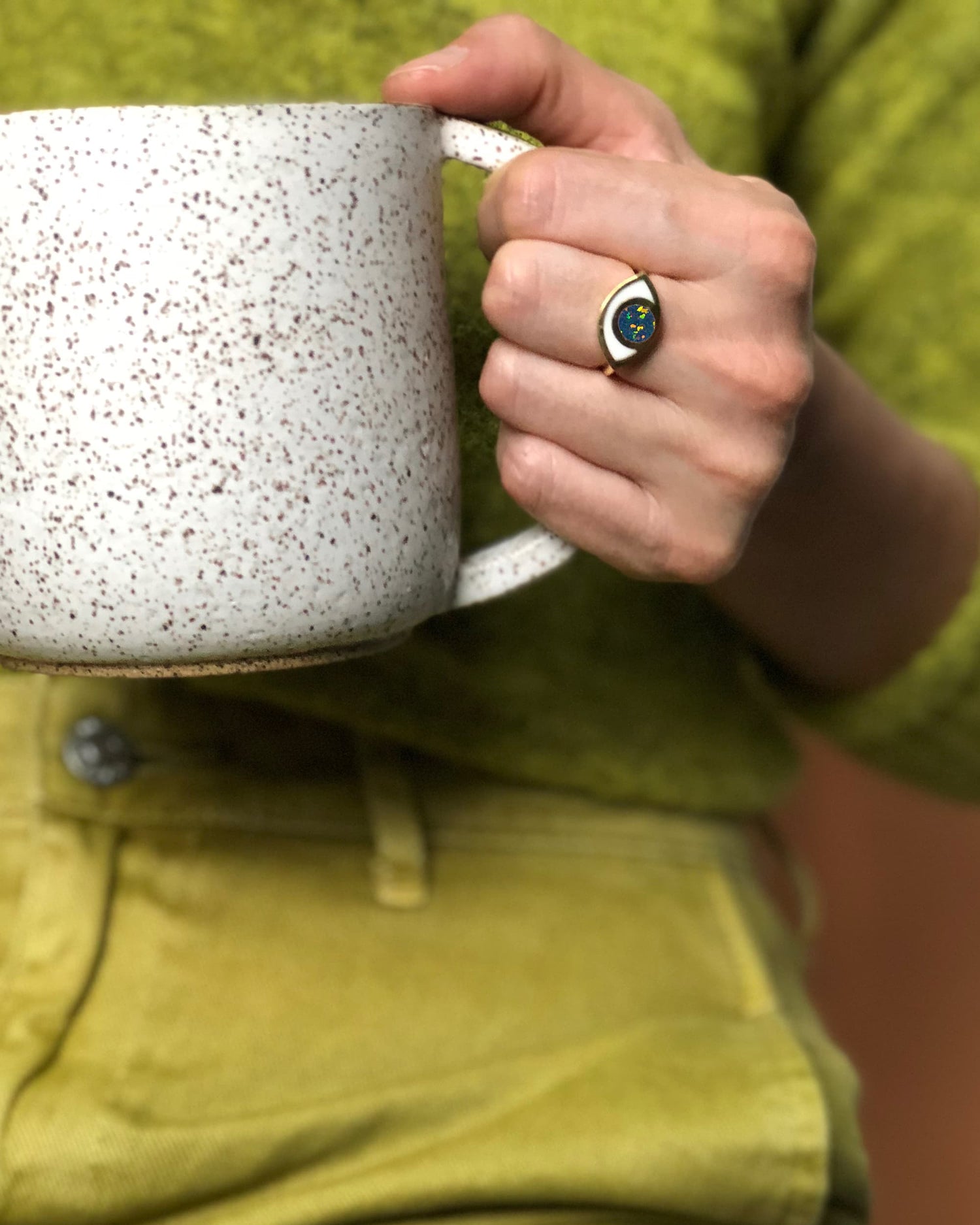 a hand wearing an eye shaped ring and holding a ceramic mug