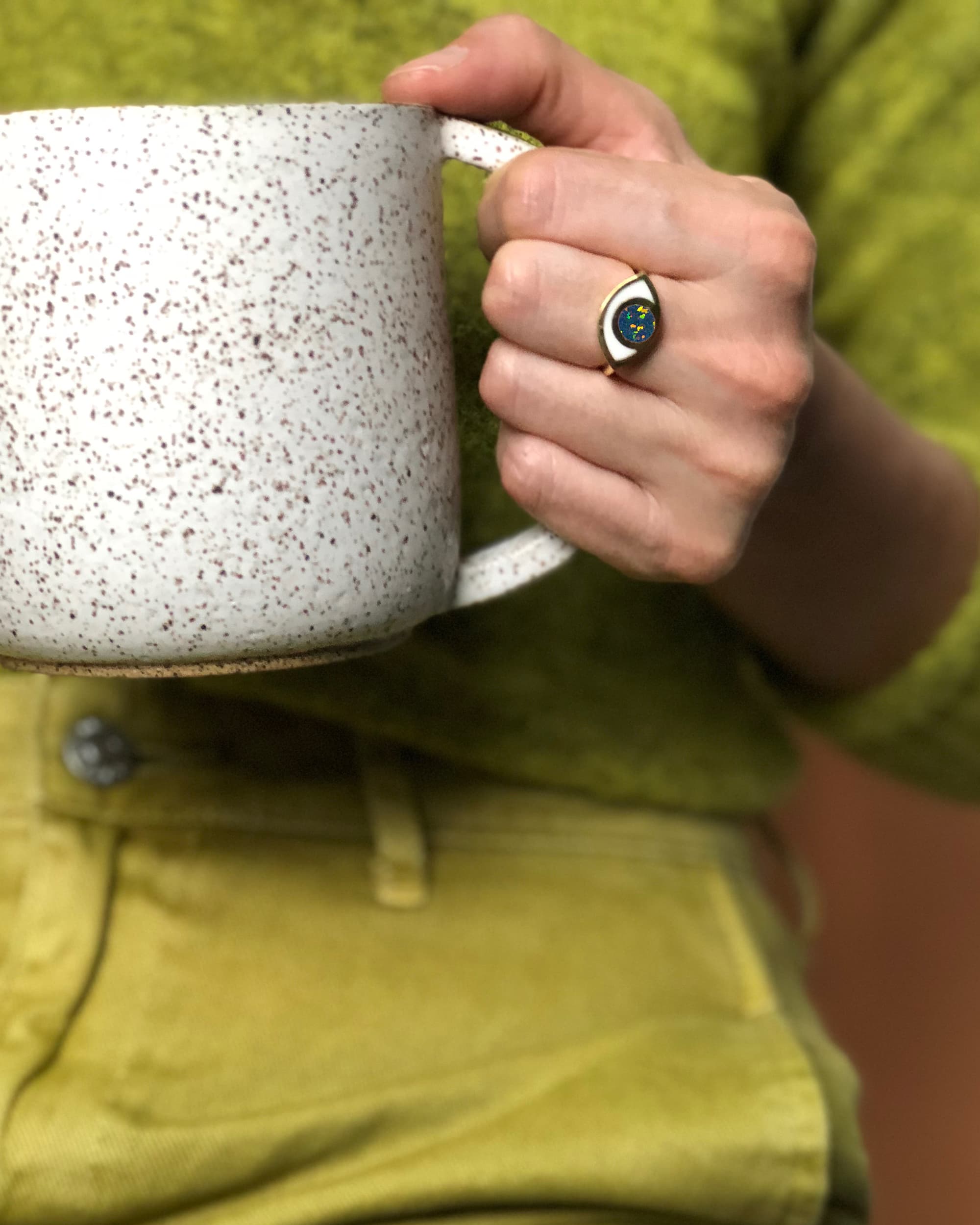 a hand wearing an eye shaped ring and holding a ceramic mug