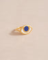 A gold ring shaped like an eye with a lapis lazuli stone set in the center, on a neutral background.