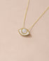 A pendant necklace with a gold eye design featuring a mother of pearl inset, on a gold chain against a beige background.