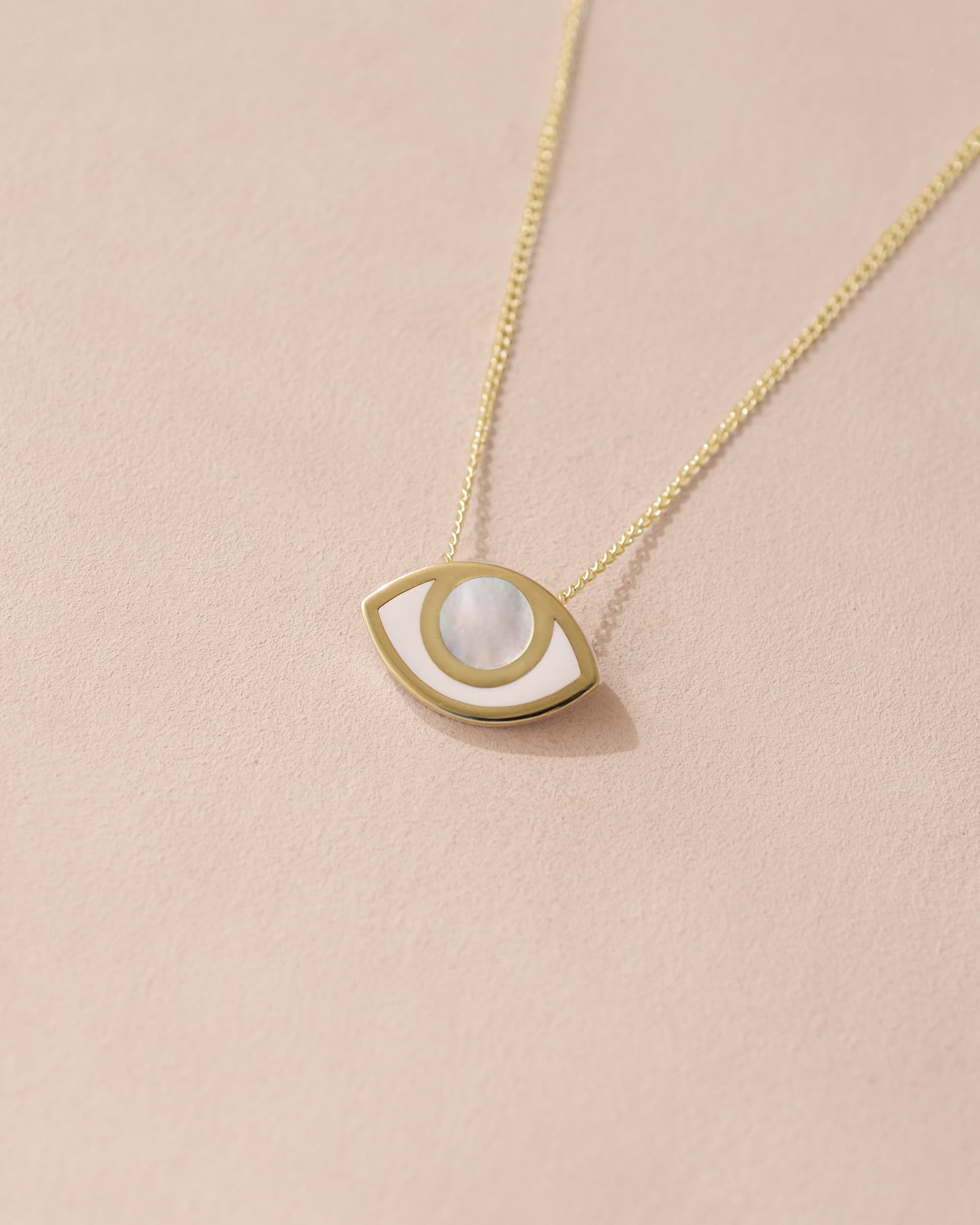 A pendant necklace with a gold eye design featuring a mother of pearl inset, on a gold chain against a beige background.