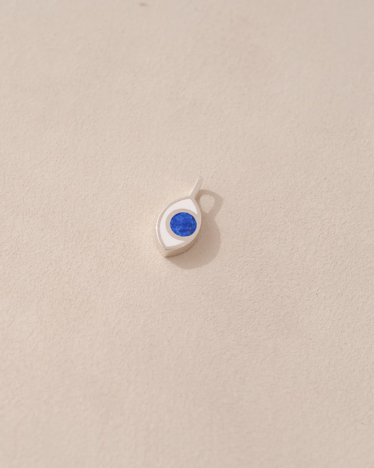 A silver charm with a lapis lazuli stone in the shape of an eye, placed against a beige background.