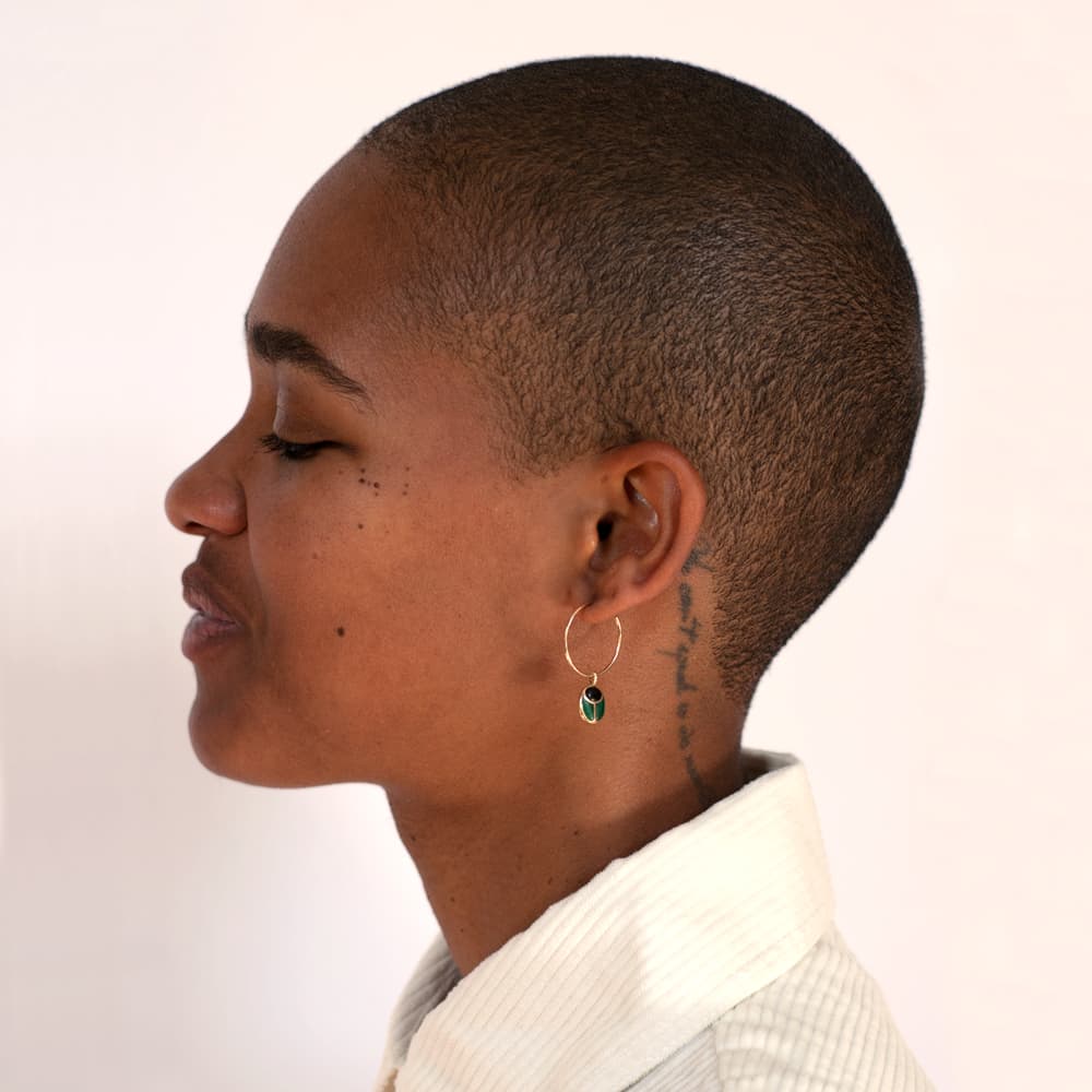 a profile view of a model wearing a scarab shaped gold charm hanging from a hoop on her ear