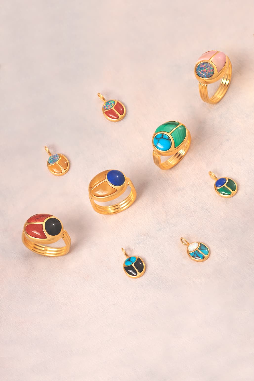 four gold rings and five gold scarab shaped charms featuring many colorful stones on a neutral background