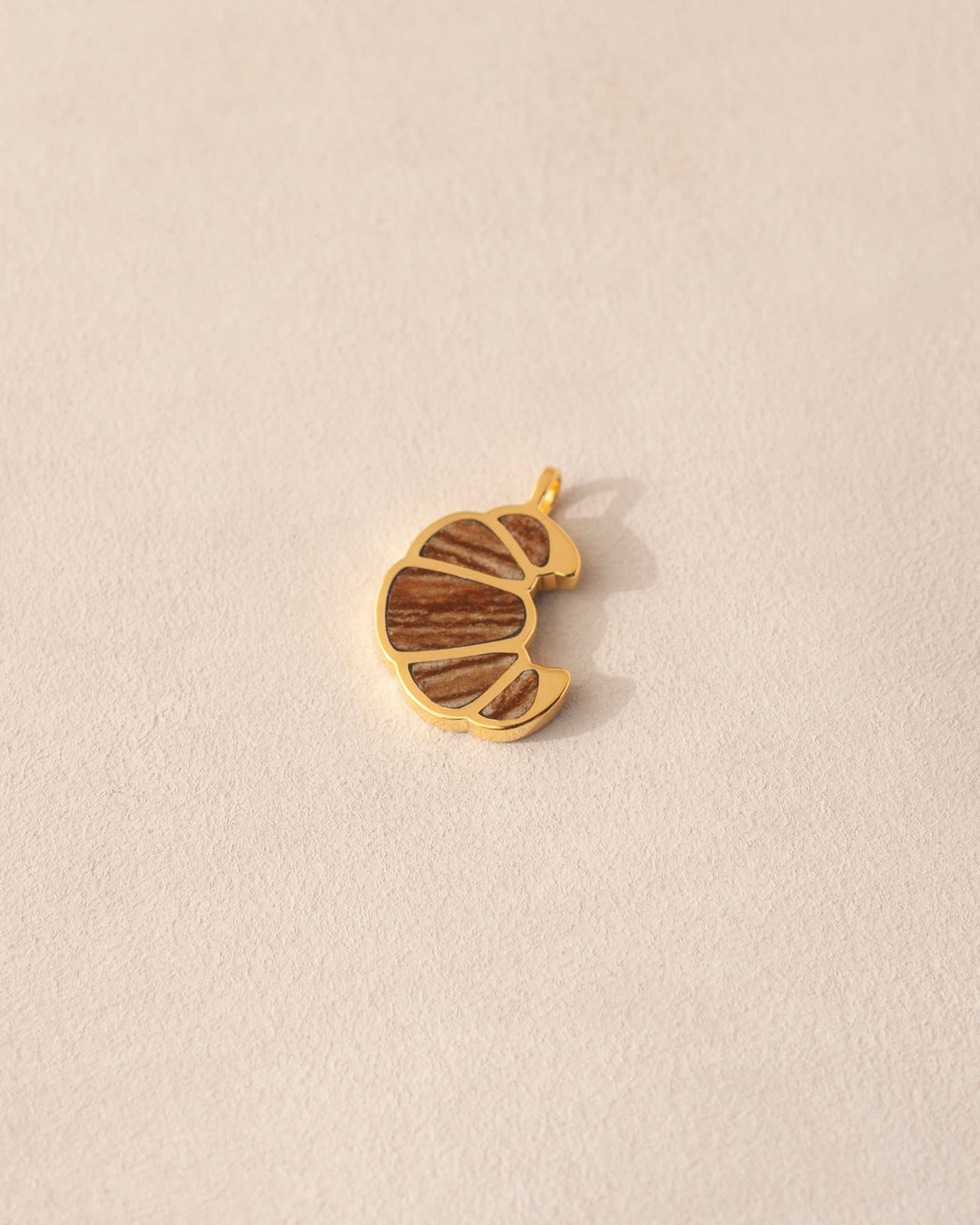 A gold charm in the shape of a croissant inlaid with jasper, displayed on a beige background.
