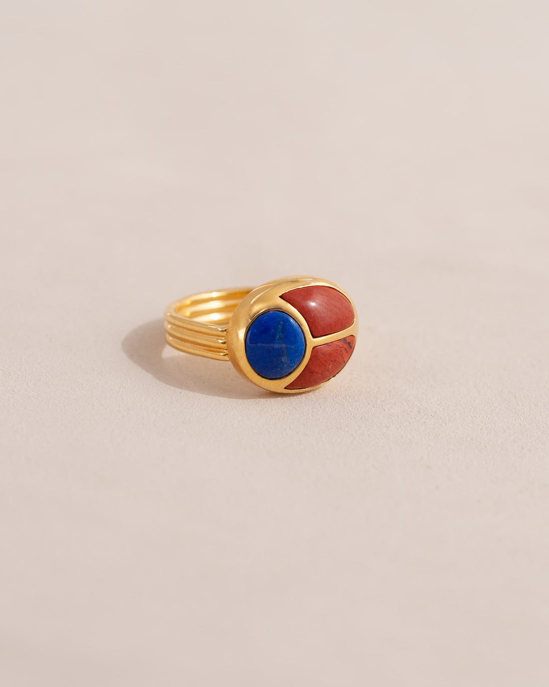 A gold ring featuring a scarab design with lapis lazuli and red jasper inlays, displayed against a neutral background.