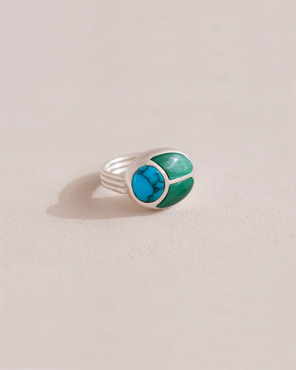 A silver ring featuring a central turquoise stone flanked by malachite stones on either side, set against a neutral background.