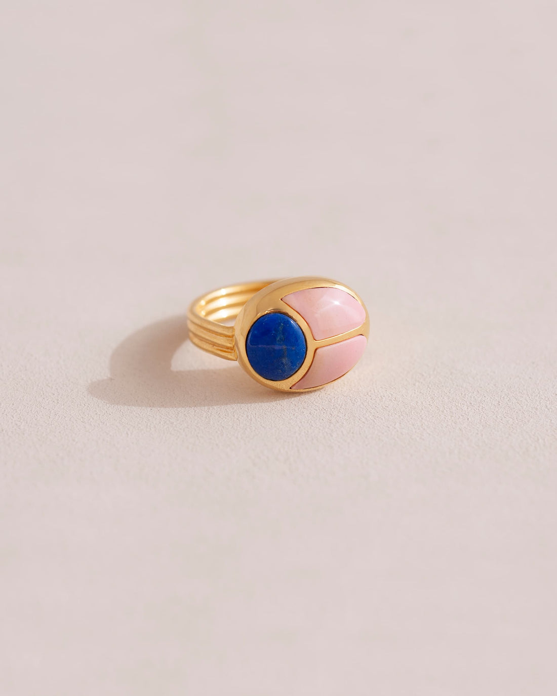 A gold ring featuring a central lapis lazuli stone flanked by pink opal stones on either side, set against a neutral background.