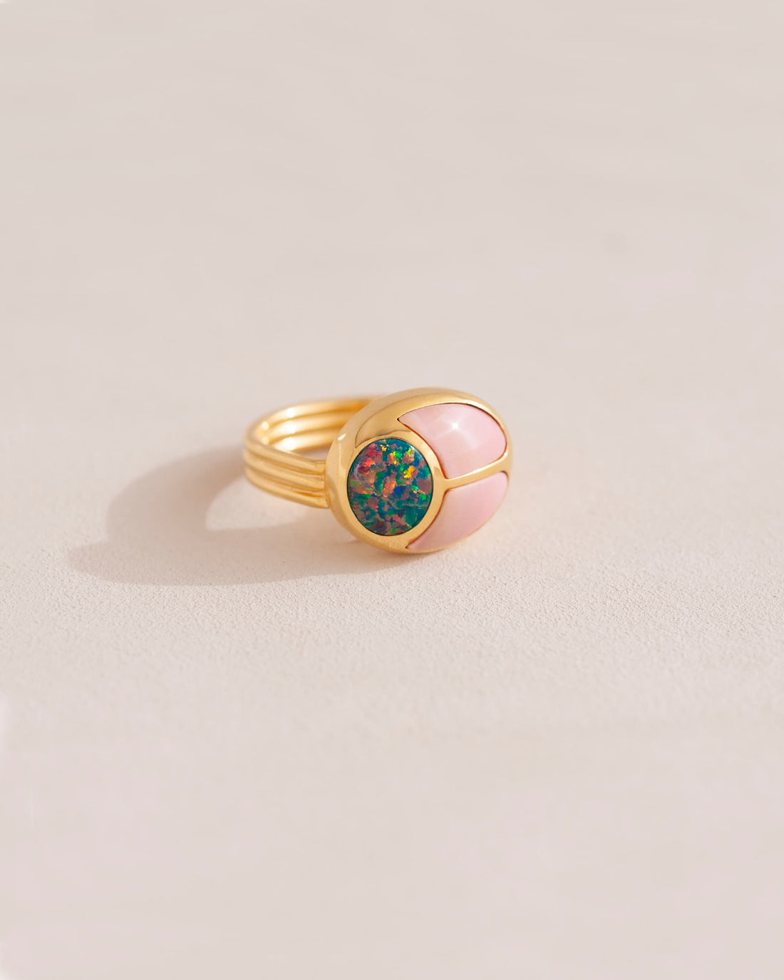 A gold ring featuring a central opal stone flanked by pink opal stones on either side, set against a neutral background.