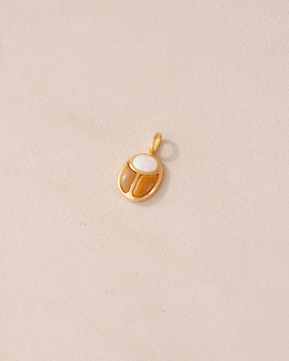 A gold-colored charm featuring a scarab design with a white opal in the center, set against a beige background.