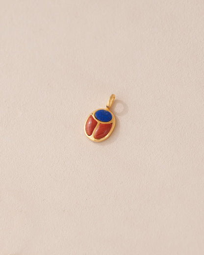 A gold charm featuring a scarab design with lapis lazuli and red jasper inlays, displayed against a neutral background.