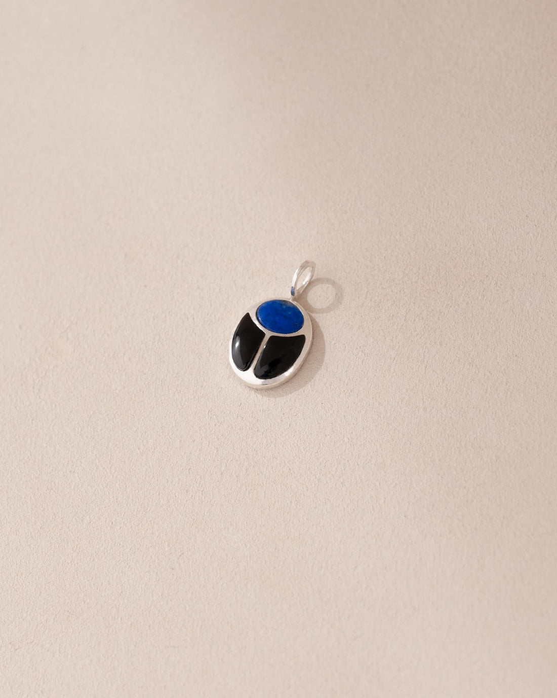 A silver-colored charm with a blue lapis lazuli center and black onyx surround, placed against a beige background.