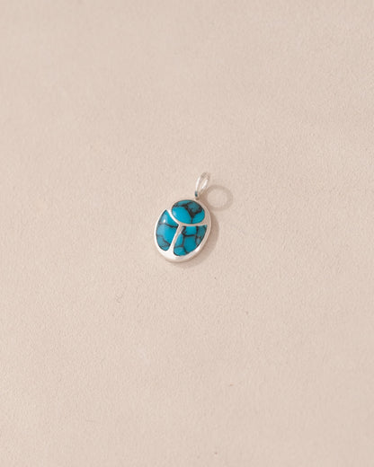 A small turquoise stone charm with a metallic setting, shaped like a scarab, displayed against a light background.