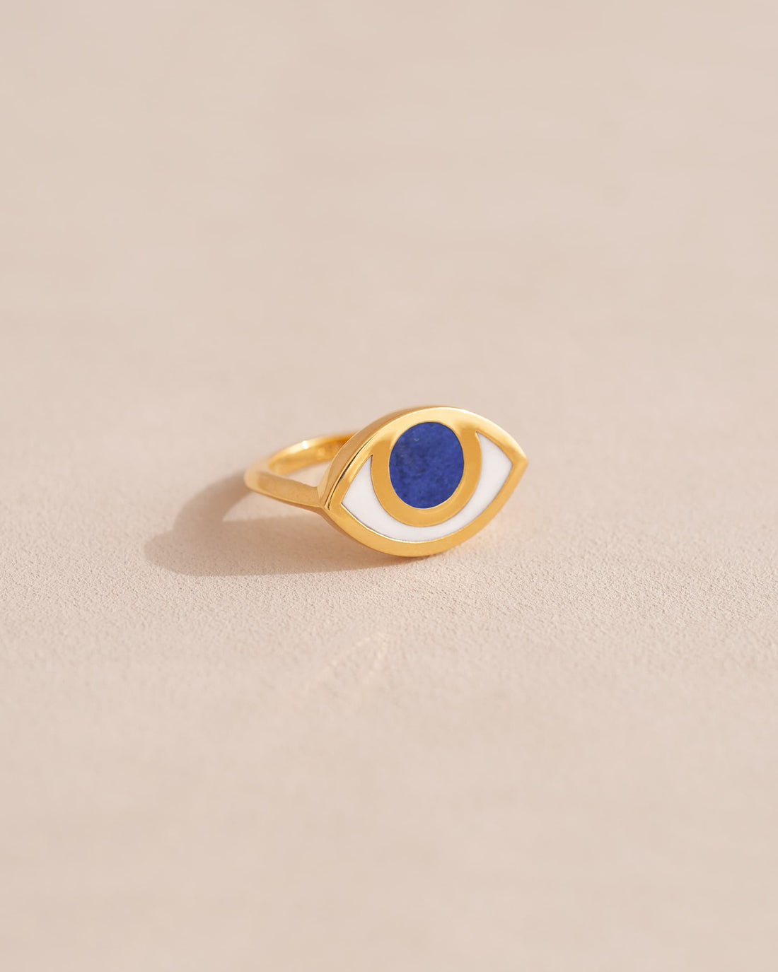 A gold ring shaped like an eye with a lapis lazuli stone set in the center, on a neutral background.