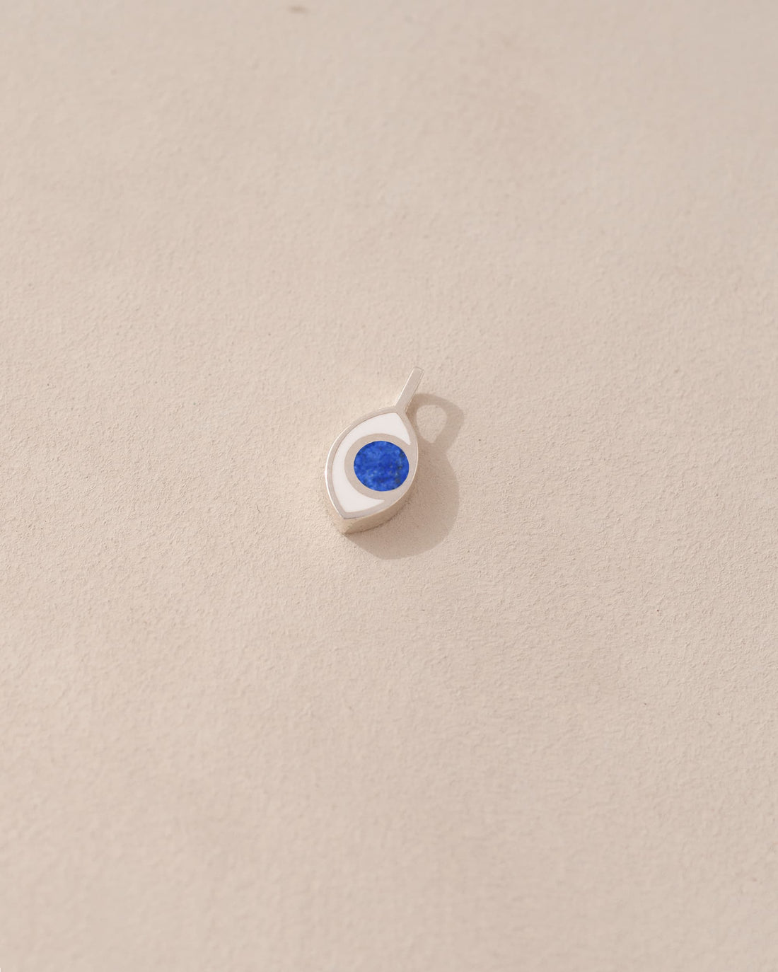A silver charm with a lapis lazuli stone in the shape of an eye, placed against a beige background.